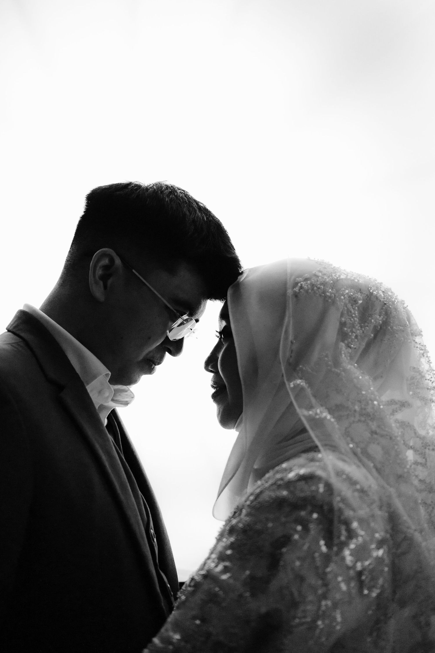 Black and white photo of a Muslim couple in wedding attire, groom in suit and bride in hijab, touching foreheads romantically.