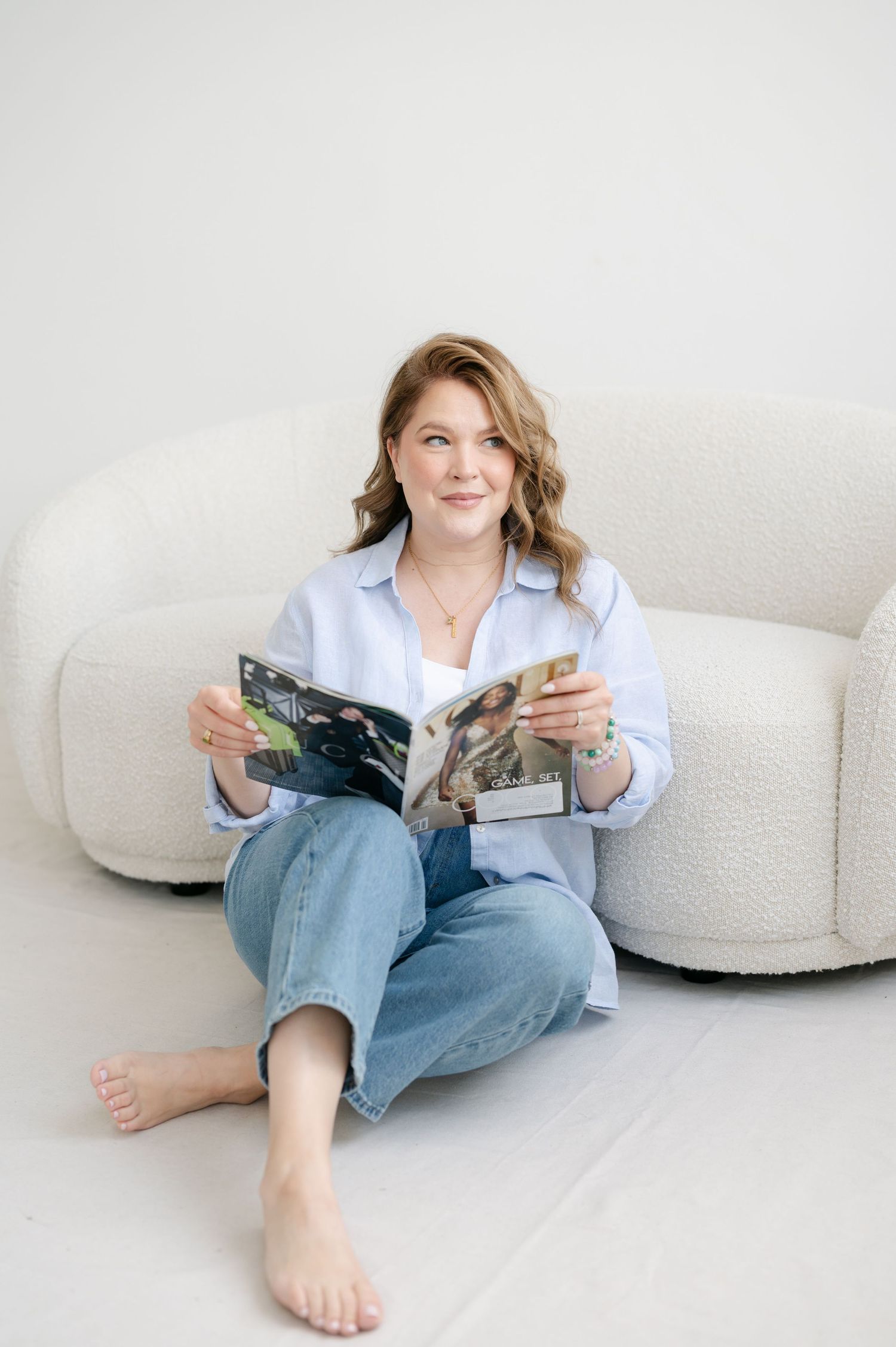 A person in a white shirt and jeans sits barefoot on the floor reading a magazine near modern curved furniture.