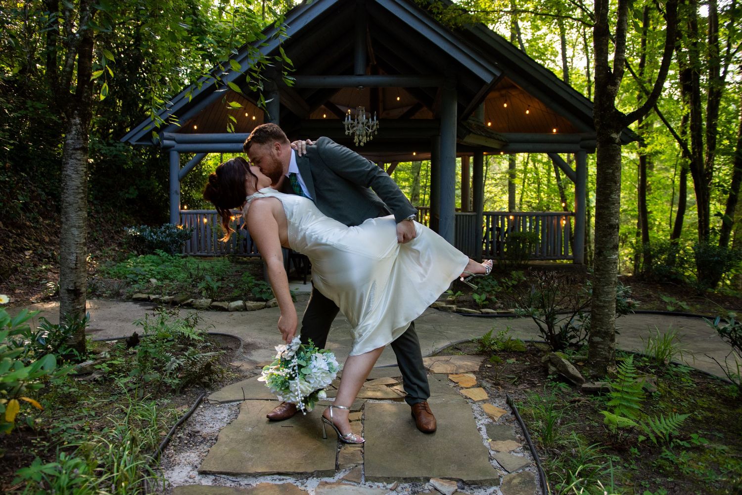 A romantic dip pose captured at a wooded outdoor wedding venue with natural sunlight filtering through trees.