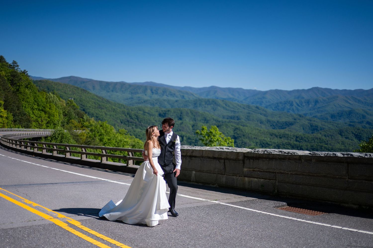 Bride and groom share a romantic kiss on a scenic mountain road with lush green hills and blue sky backdrop.