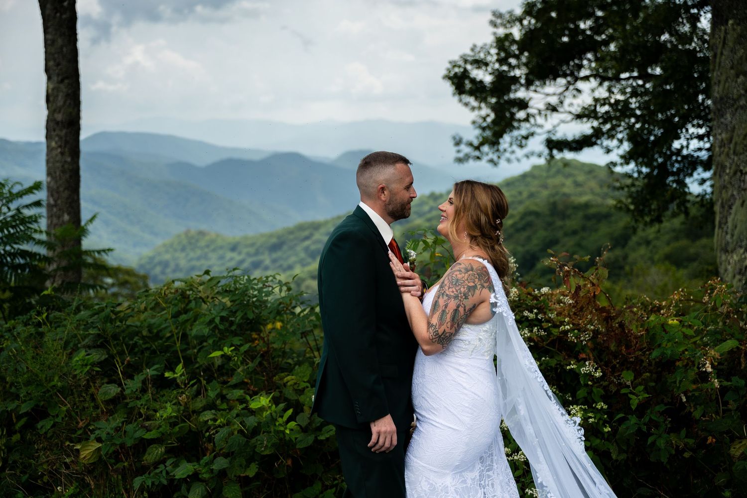 Bride in a white gown with a flowing veil and groom in a dark suit share an intimate moment overlooking misty mountains at a scenic wedding.