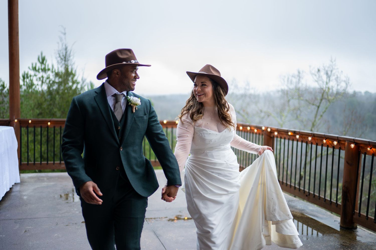 Bride and groom in cowboy hats hold hands walking on rainy outdoor deck at rustic wedding venue.