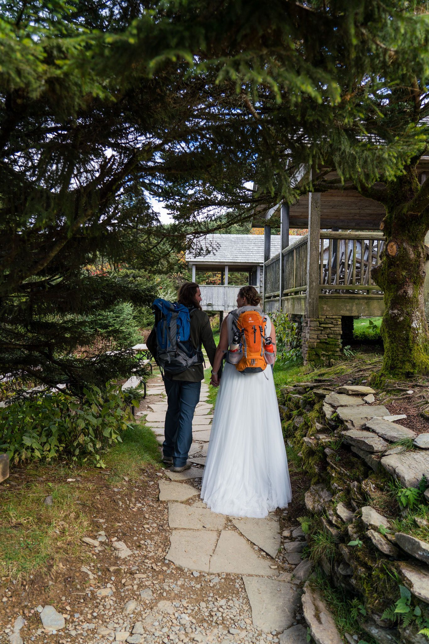 Bride in white dress and groom with hiking backpacks hold hands on stone path near rustic cabin surrounded by trees.
