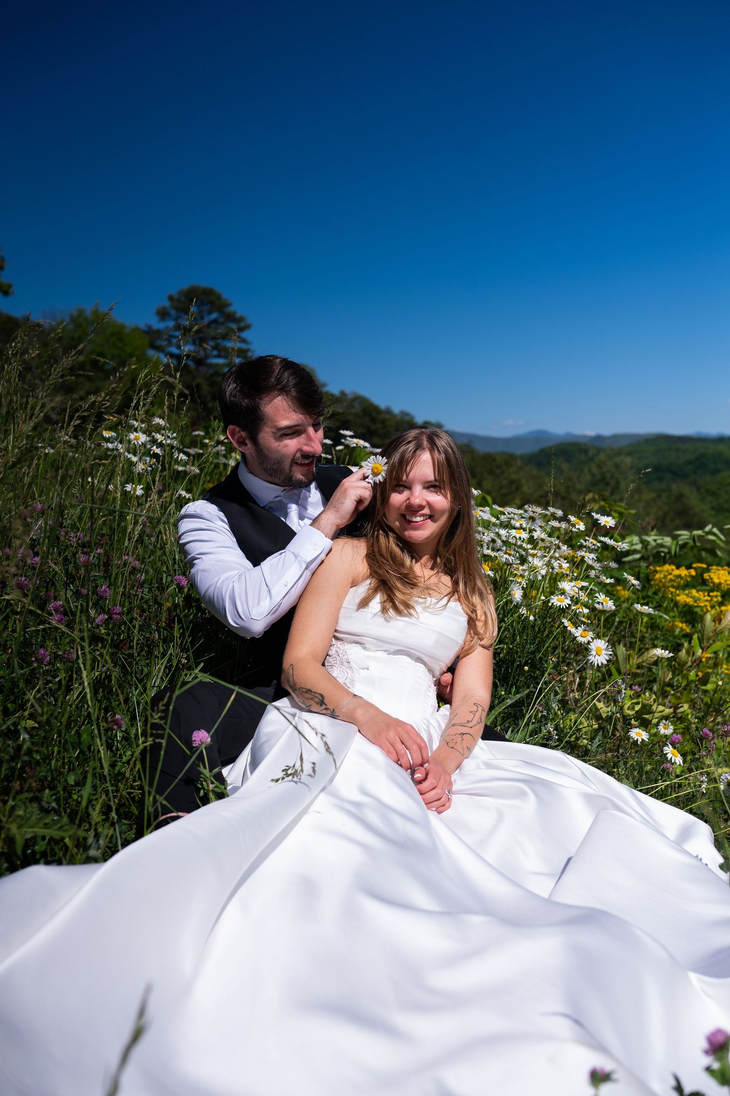 Bride and groom sitting in a wildflower meadow on a sunny day, with mountains and blue sky in the background.
