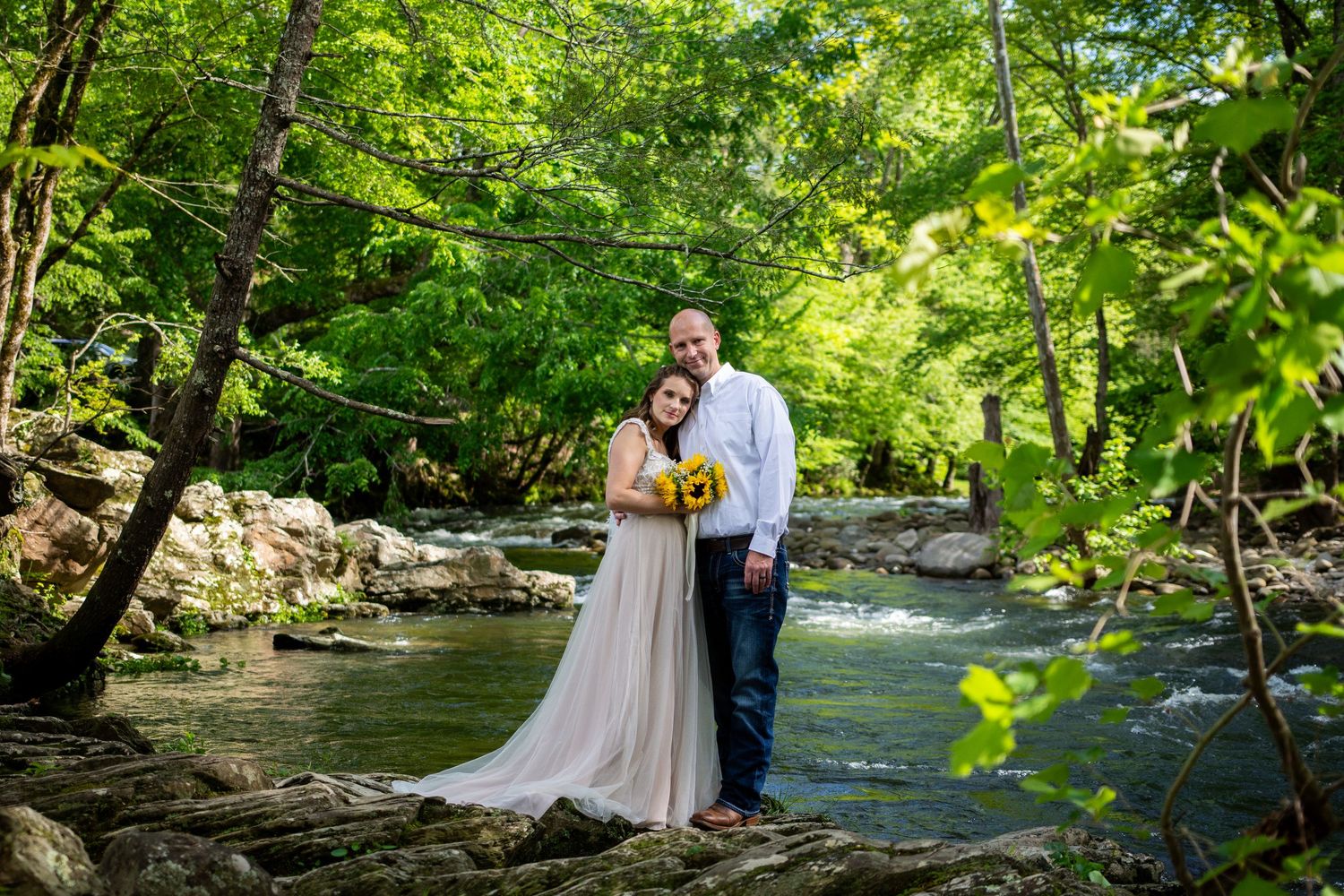 A couple in wedding attire poses along a scenic creek surrounded by lush green forest on their special day.