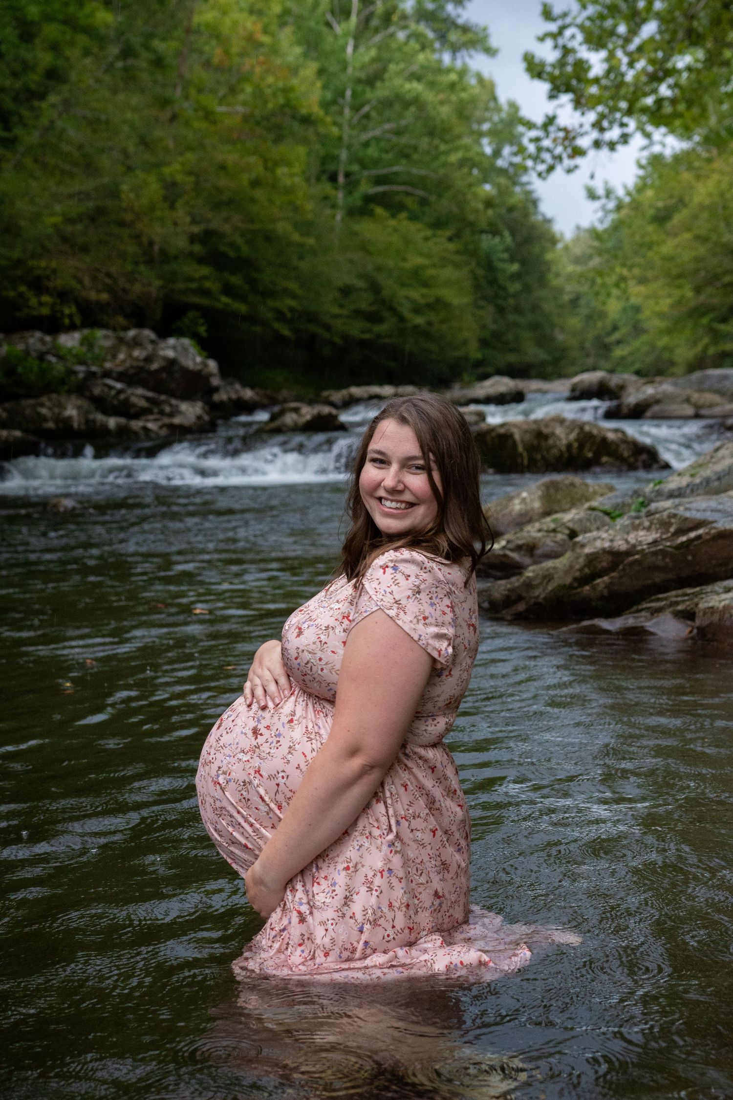 A person in a pink lace dress poses for photos in a scenic river surrounded by rocks and forest trees.