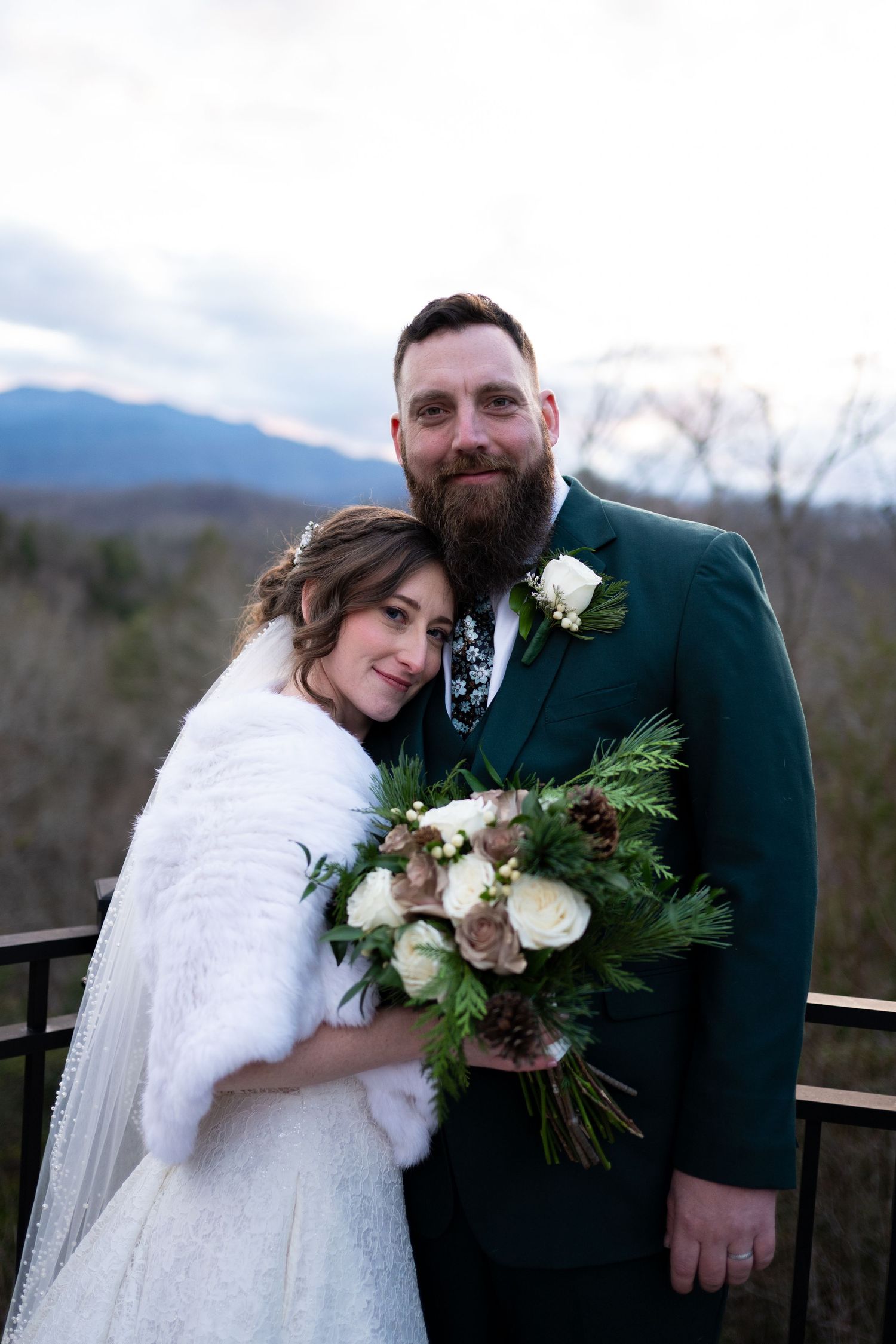 A winter wedding couple poses together on a mountain deck with evergreen bouquet accents and a white fur wrap.