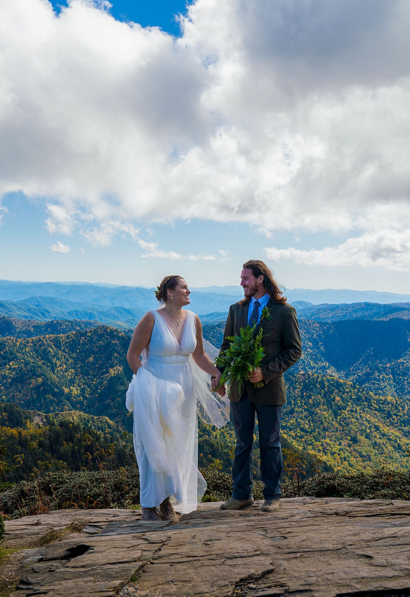 Couple in wedding attire holding hands on a mountain overlook with sweeping blue ridge mountain views under a cloudy sky.