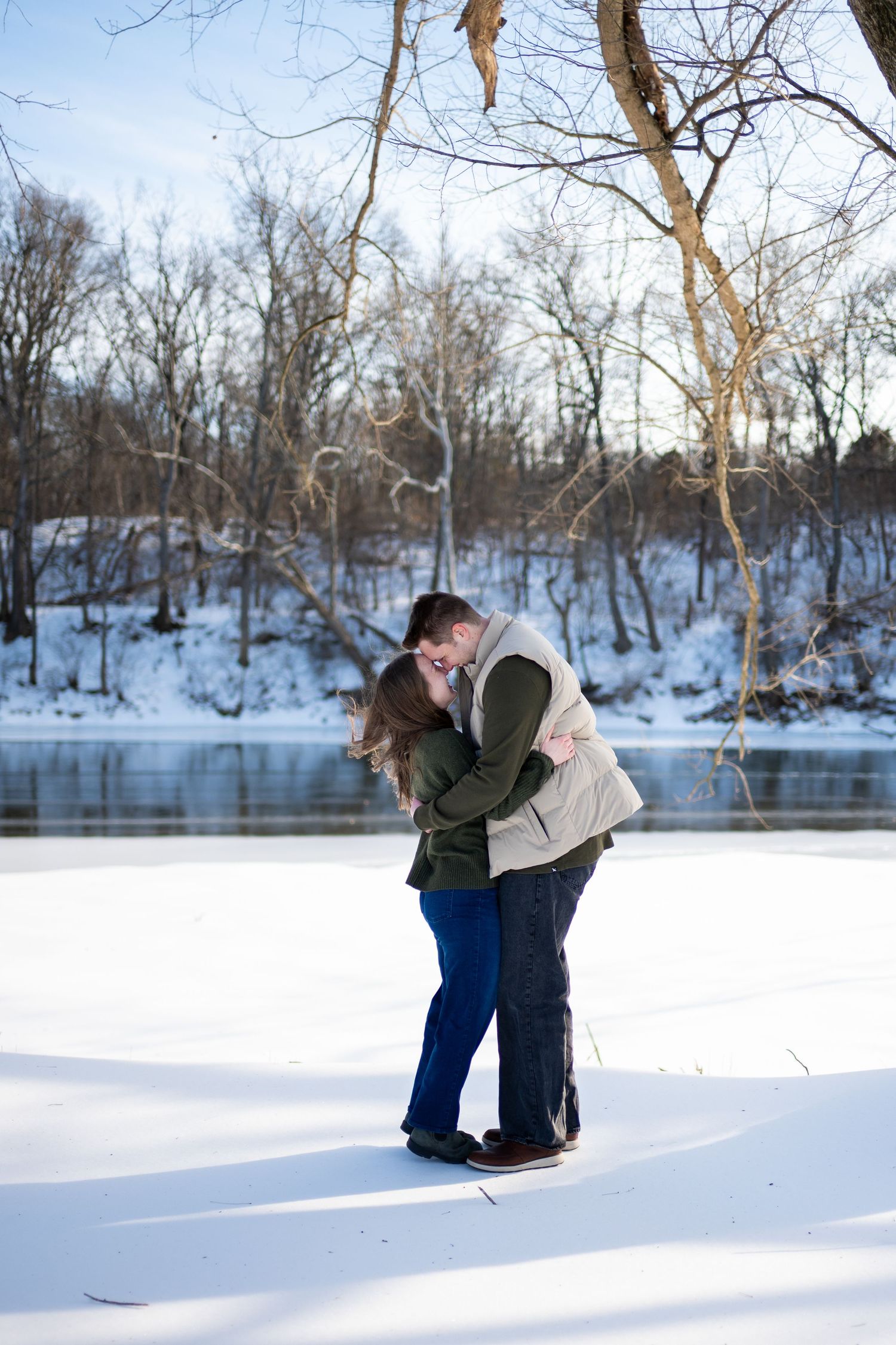 A couple embraces in a snowy winter landscape beside a frozen lake with bare trees in the background.
