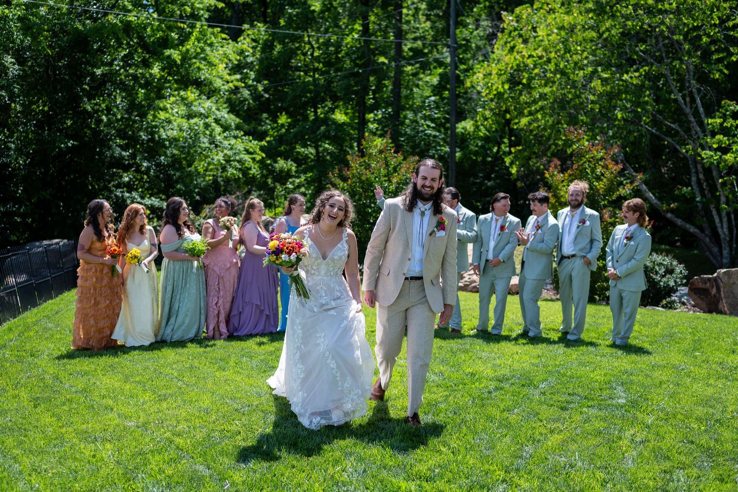 Bride and groom walk hand-in-hand on green lawn with colorful bridesmaids and groomsmen in sage suits behind them.