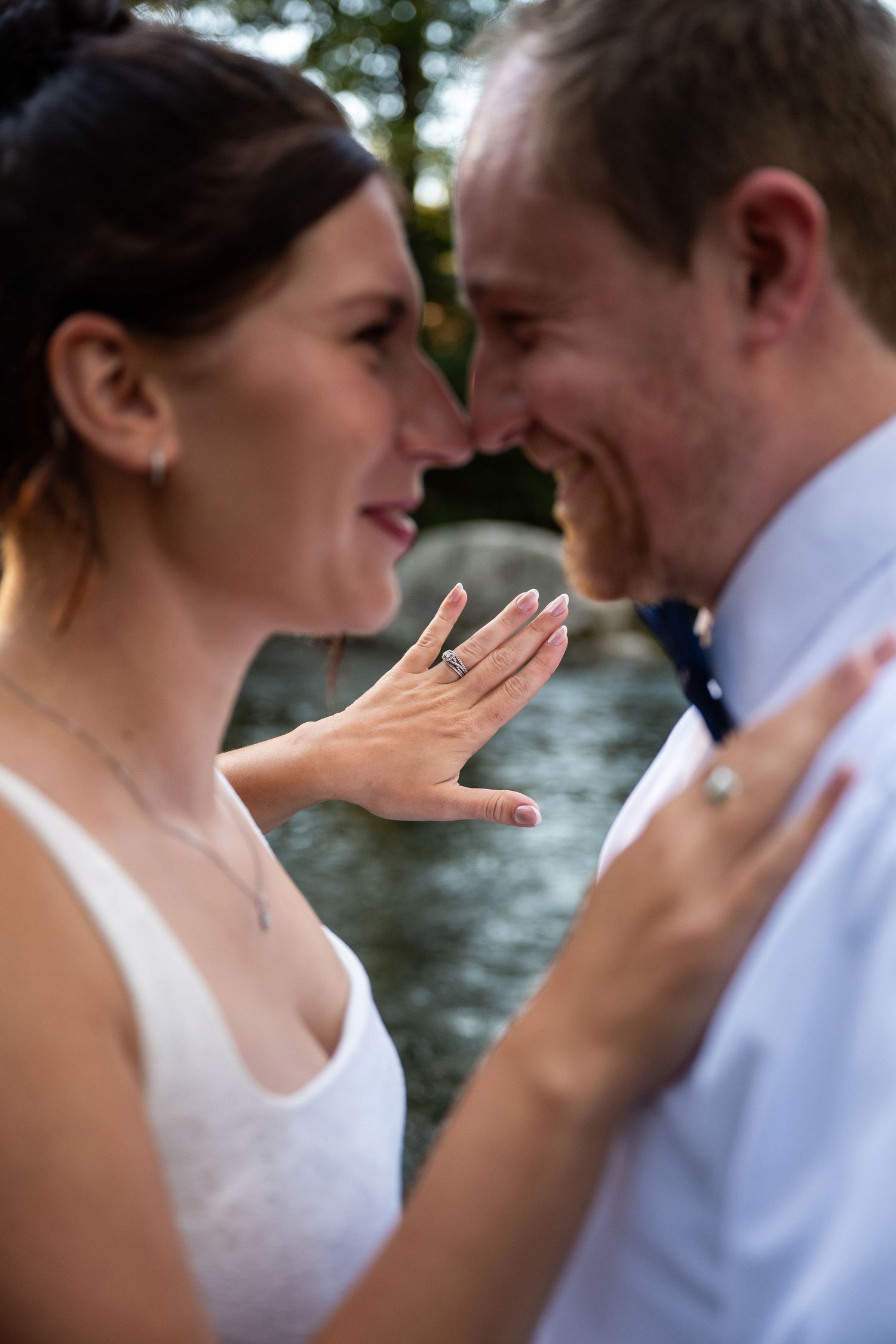 Smiling couple standing nose to nose beside a river as the woman shows off her ring on her hand resting on his shoulder.
