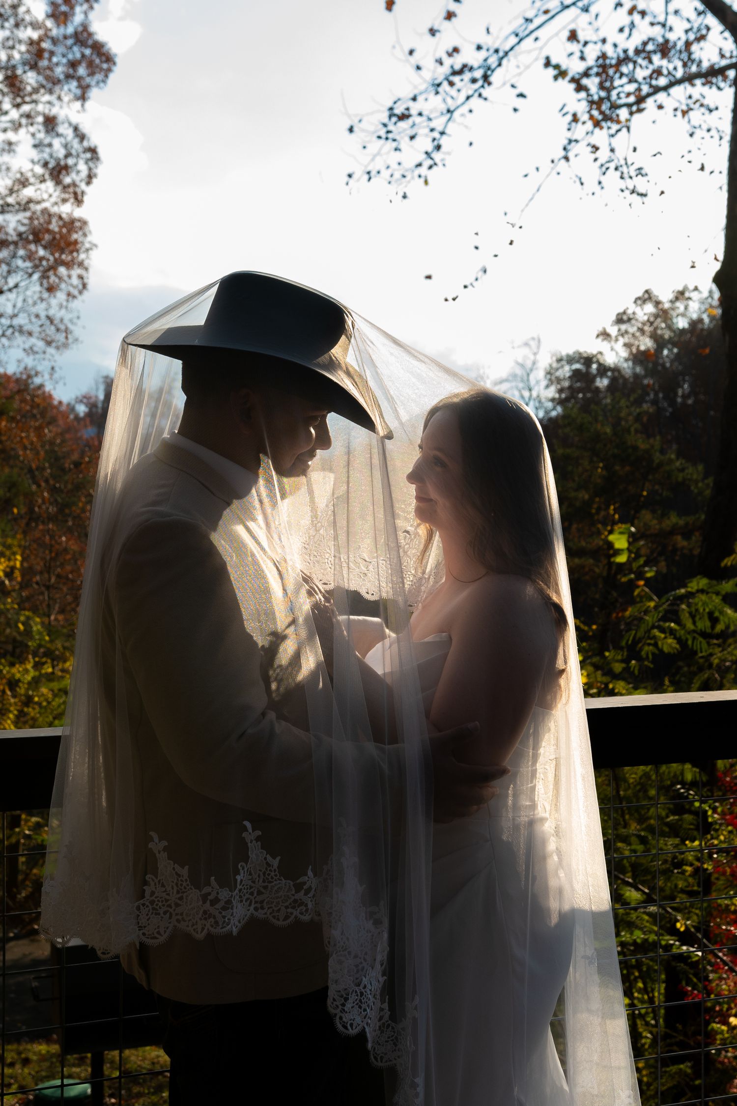 Bride and groom share an intimate moment under a wedding veil outdoors, groom wearing a cowboy hat.