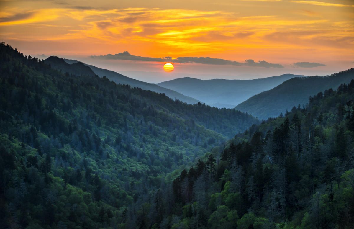 Panoramic sunset view of the Great Smoky Mountains with layered ridges silhouetted against an orange and purple sky.