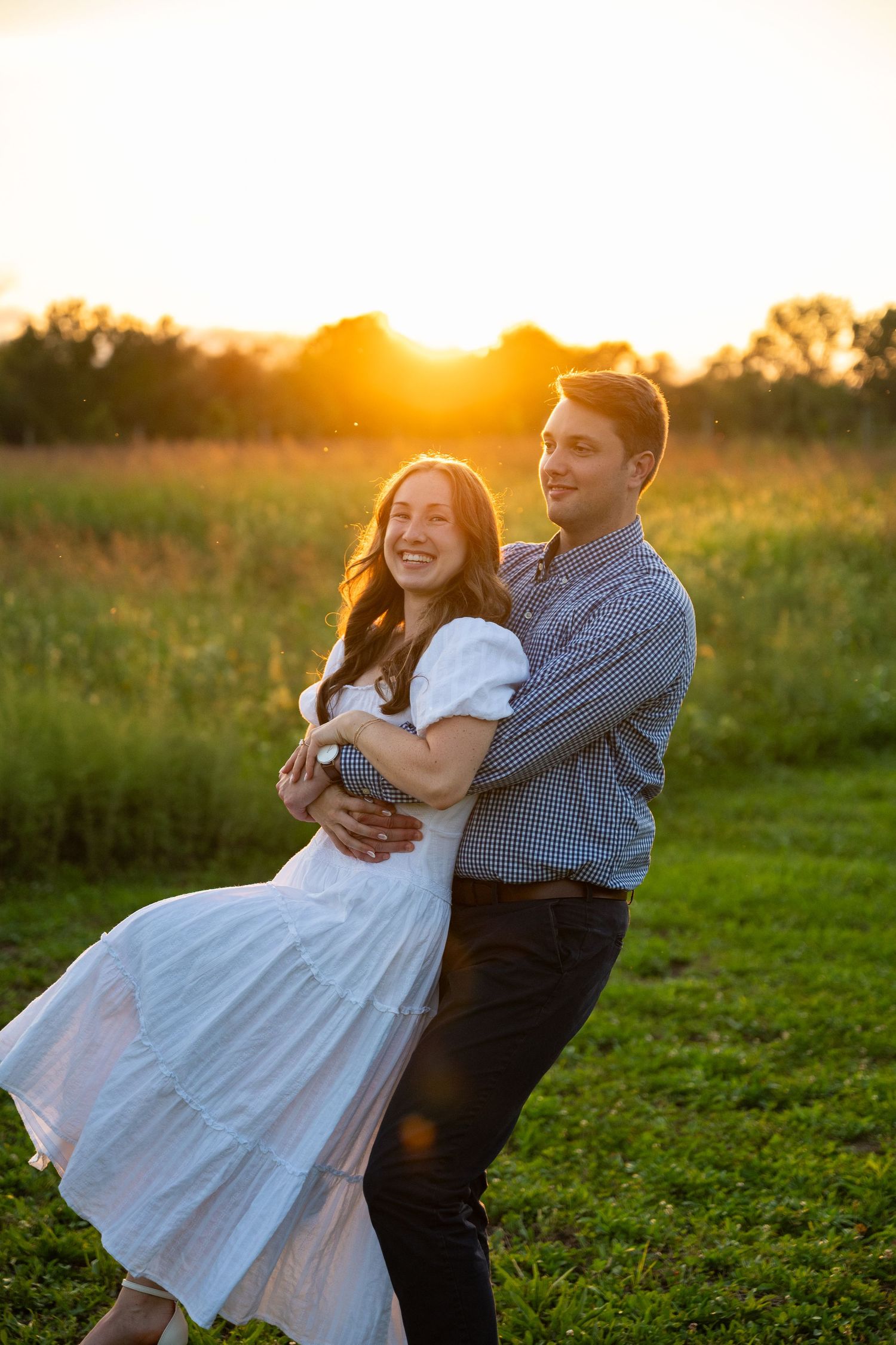 Smiling couple embracing in a grassy field at golden hour sunset, the woman in a white dress lifted playfully by her partner.