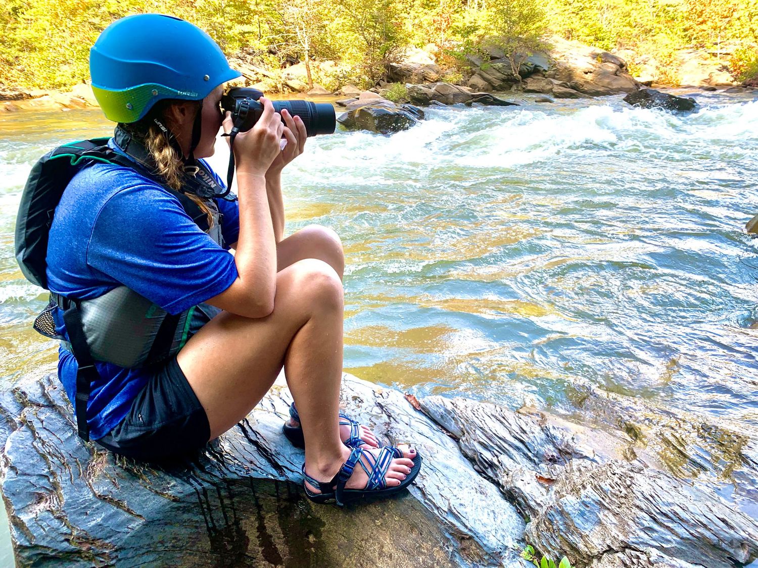 A hiker in a blue helmet sits on rocks by a rushing river, photographing the scenic waterfall landscape.