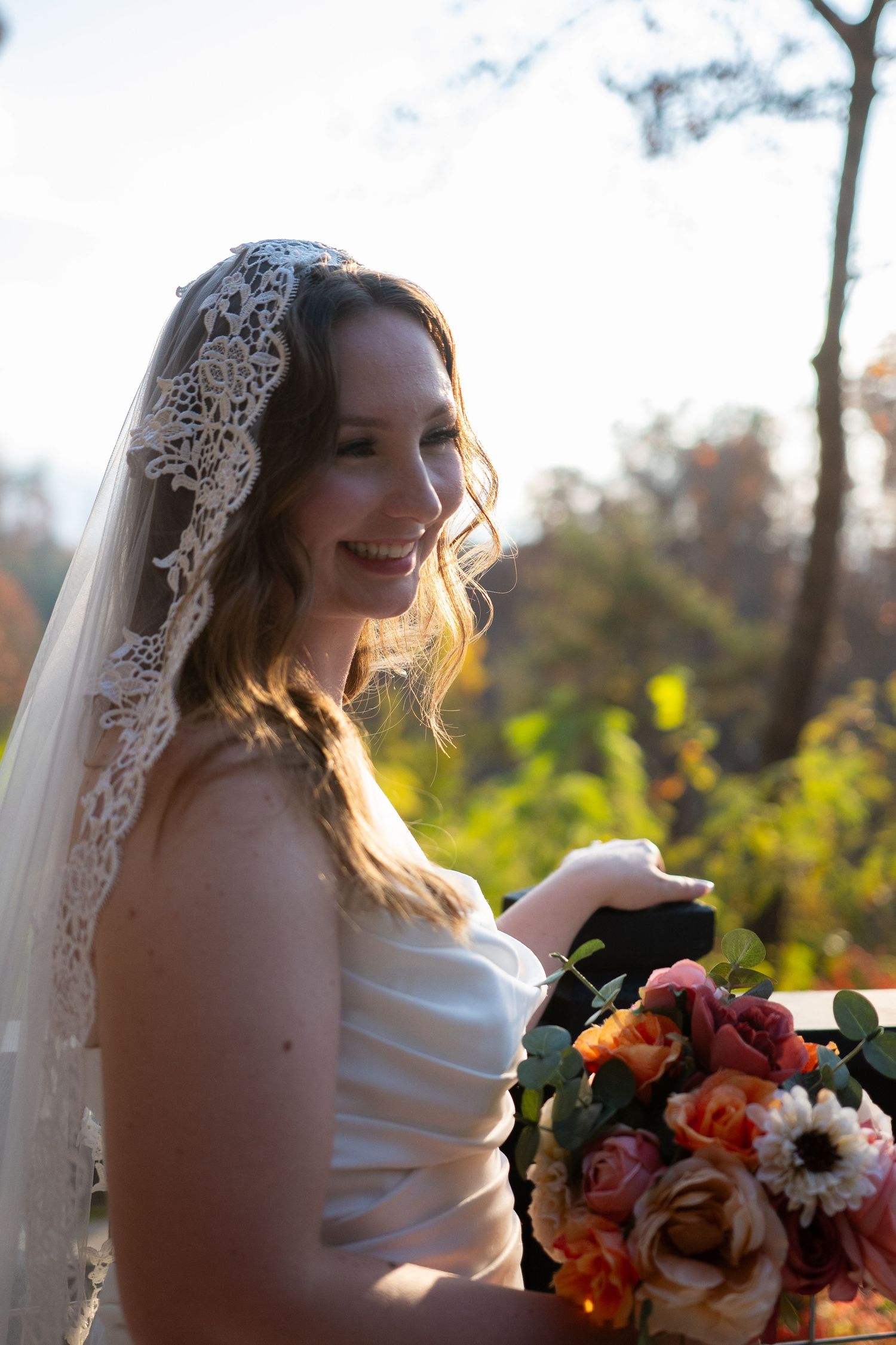 Bride in white dress with lace veil holds colorful autumn bouquet while smiling during outdoor sunset photo session.