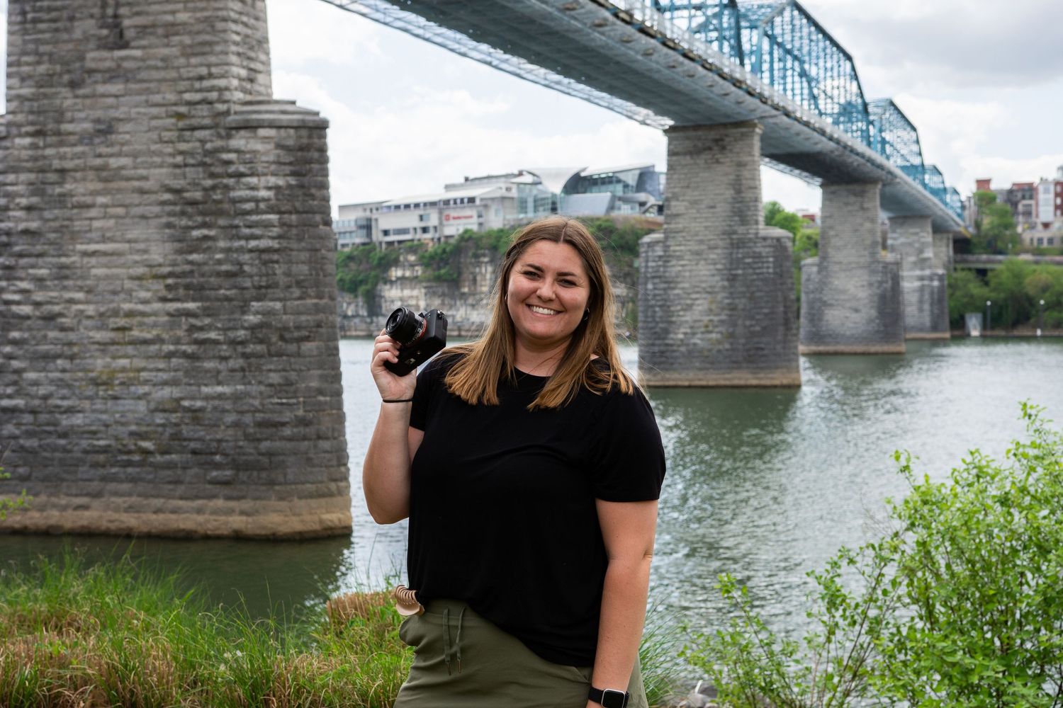 Young woman smiling and holding a camera by a river with a blue bridge in the background on a cloudy day.