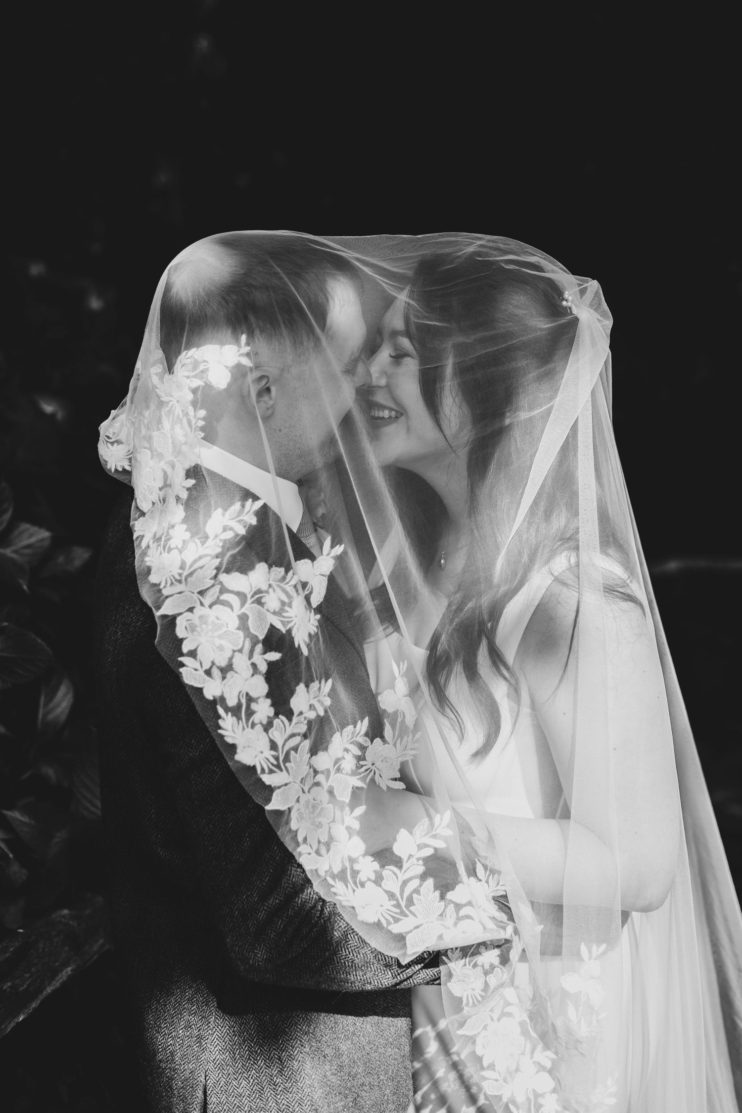 Black and white photo of bride and groom sharing a romantic moment under a lace floral wedding veil.