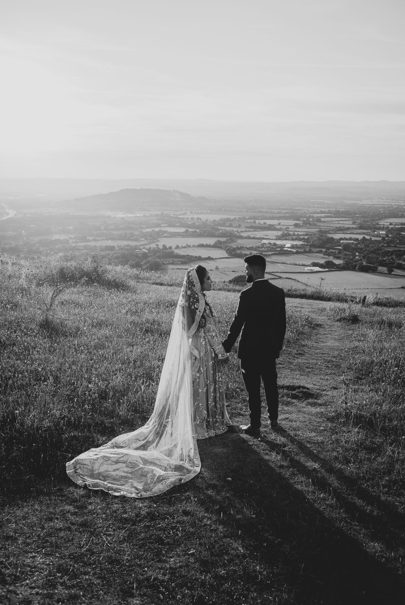 A wedding couple stands against a scenic mountain landscape with a flowing white veil in black and white photography in Cheltenham.
