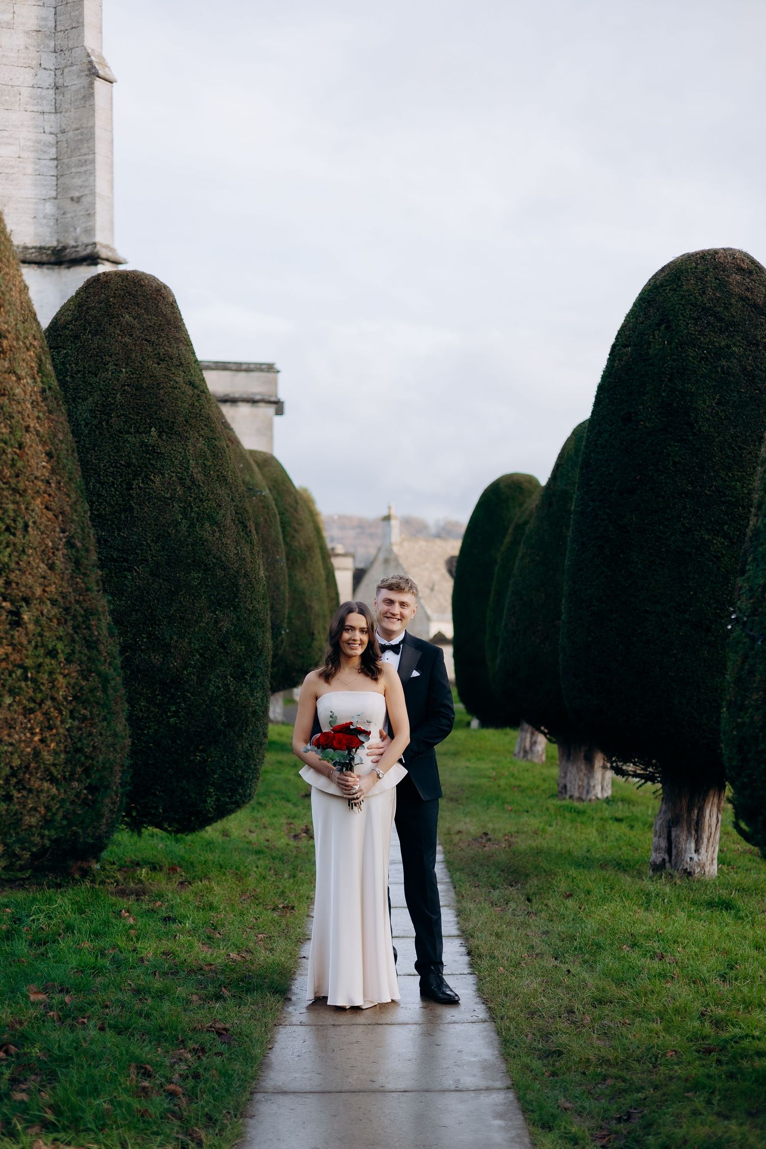 A couple poses on a garden path between tall manicured hedges during their wedding ceremony.
