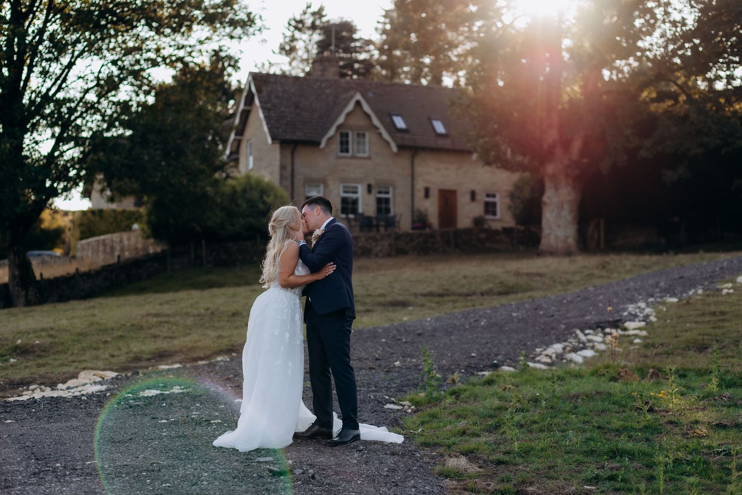 A wedding couple having their wedding portraits at kingscote barn during sunset with a stone house and garden in the background.