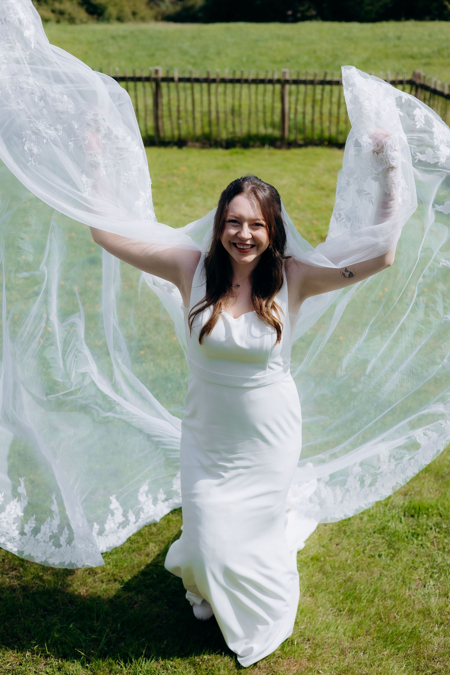 Smiling bride in white gown holding lace veil wide open outdoors on green lawn on sunny day.
