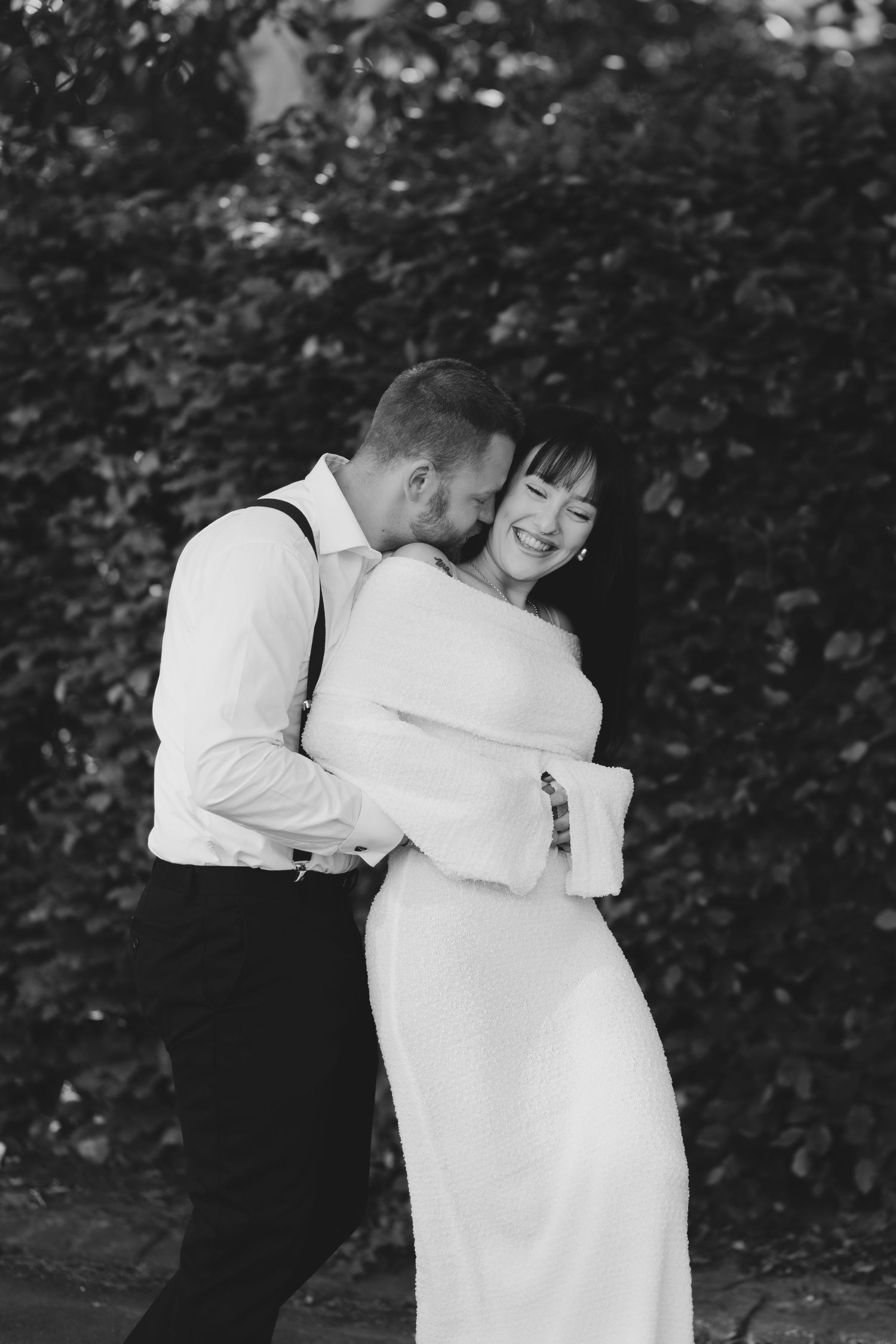 A romantic black and white cotswolds wedding photograph showing a couple embracing outdoors against a leafy backdrop.