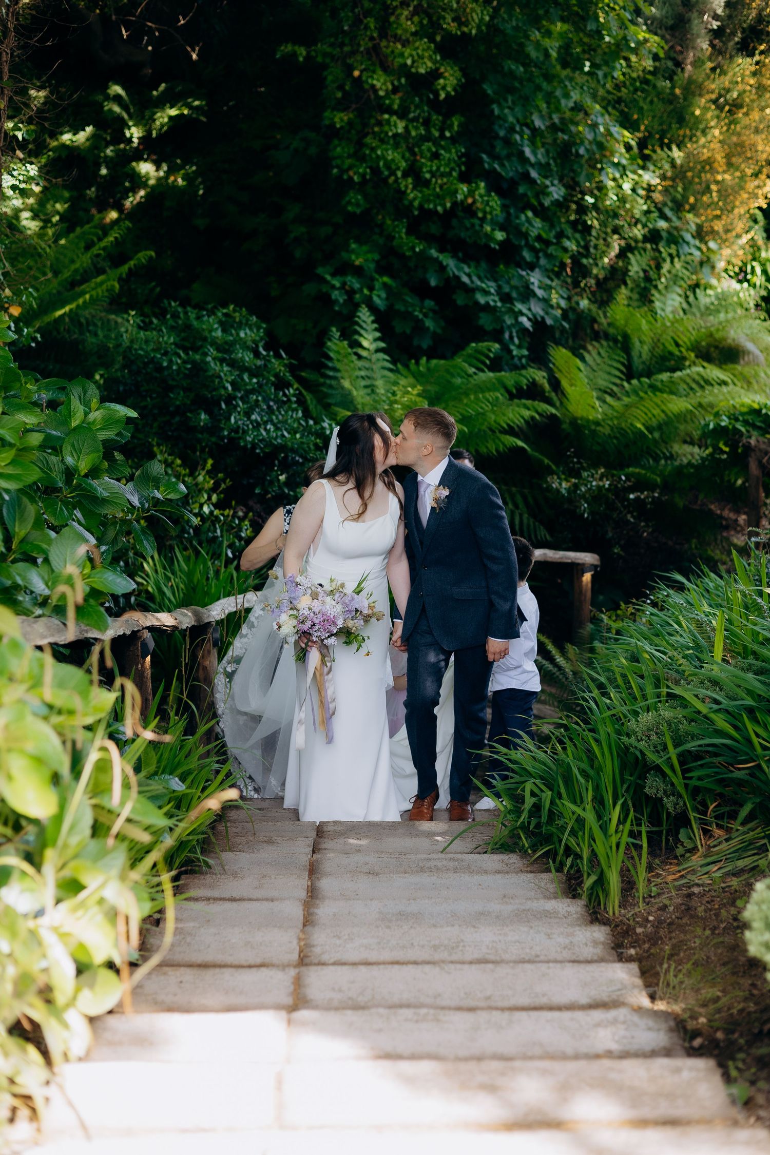 Bride and groom share a kiss on garden stone steps surrounded by lush green ferns and foliage, bride holding purple bouquet.