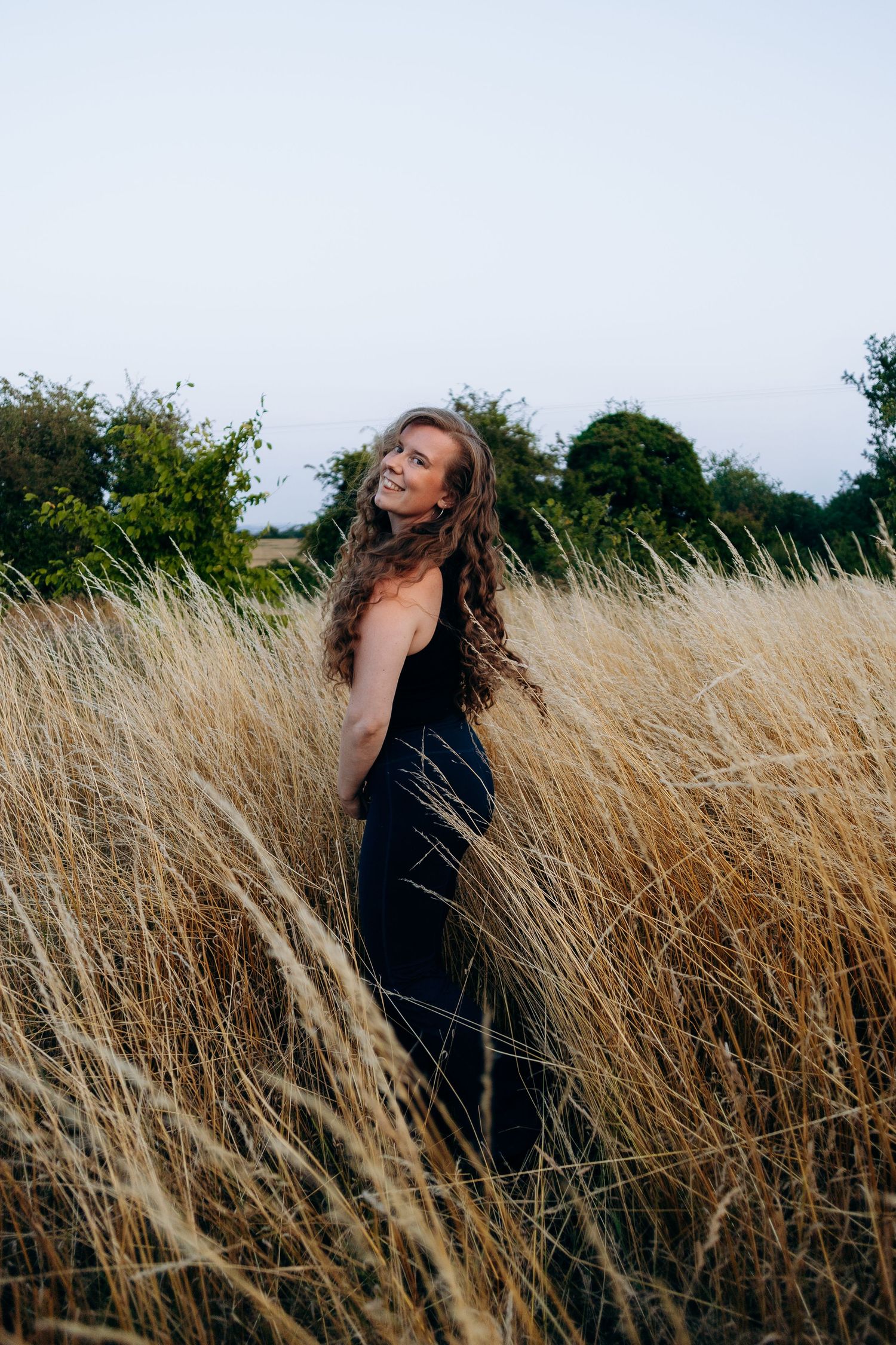 A figure in a black dress stands gracefully among tall golden wheat fields swaying in the breeze.