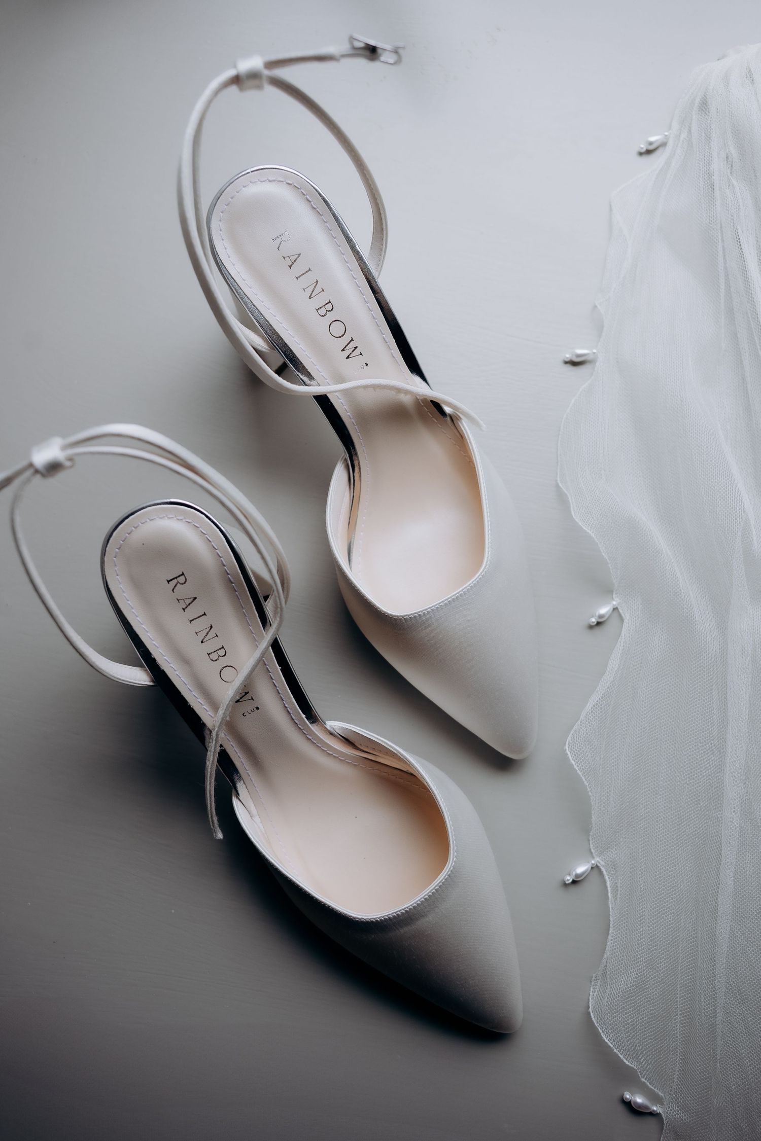 Elegant white slingback bridal shoes photographed next to sheer tulle veil on reflective surface in the cotswolds.