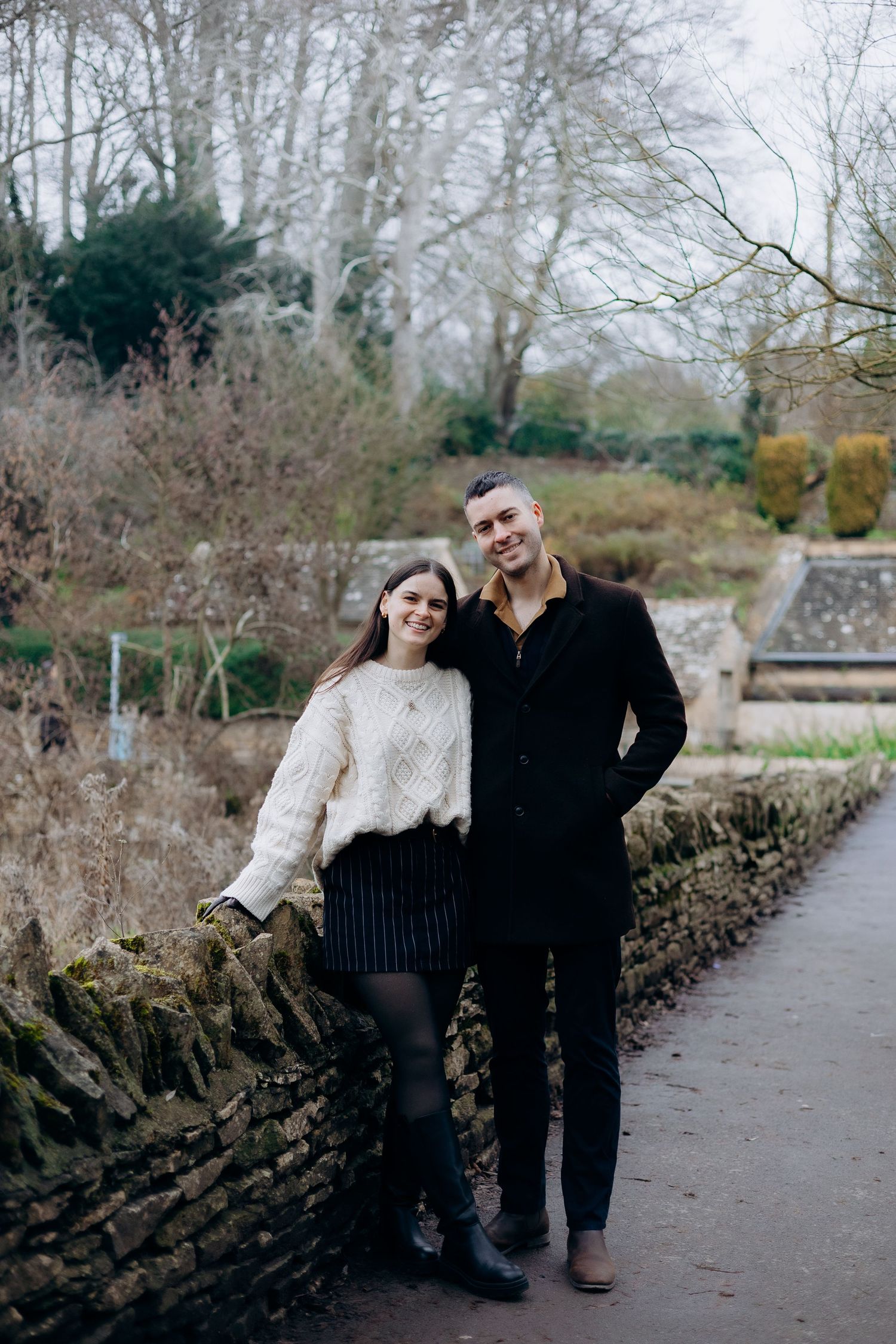 A couple shares a romantic moment walking together along a stone-lined garden path on an overcast winter day.
