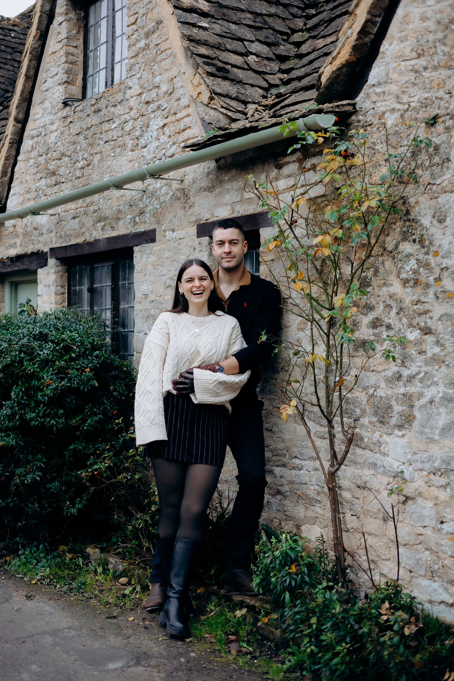 A couple stands together against a rustic stone wall with greenery in a series of romantic outdoor portraits.