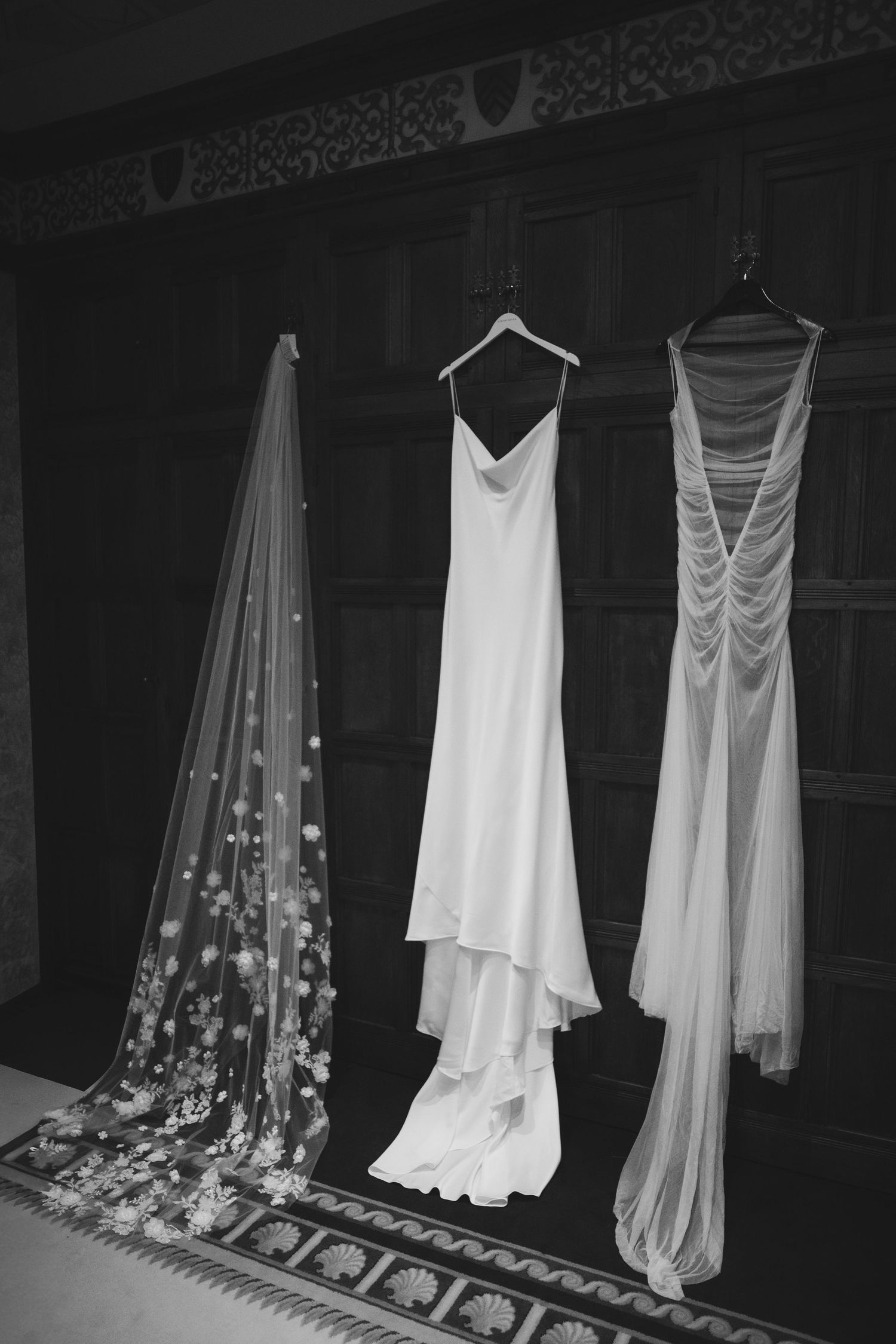Three elegant white wedding dresses and a floral veil hanging on display against a dark wooden wall in black and white at thornbury castle.