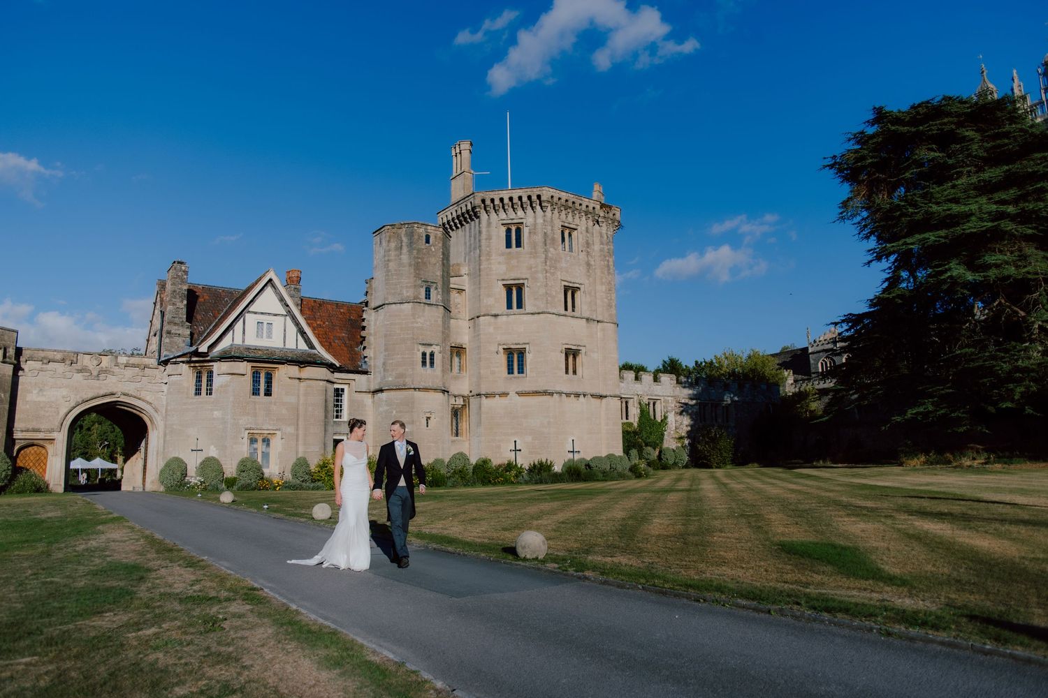A scenic wedding venue with historic castle and chapel buildings nestled among green lawns and trees on a sunny day.