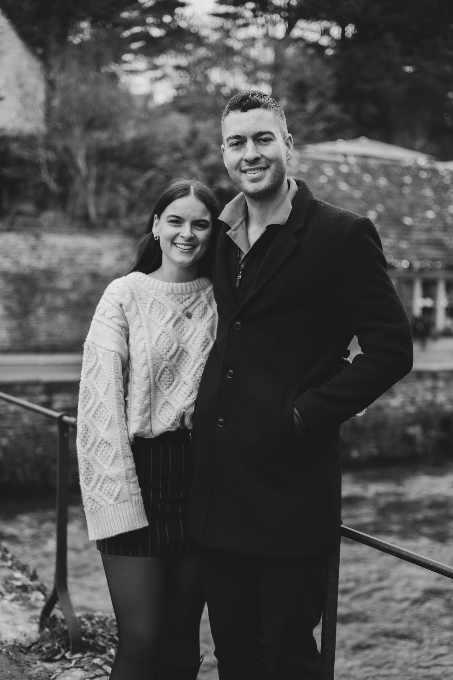 A black and white portrait of a couple outdoors, with one wearing a cable knit sweater and the other in a dark coat.