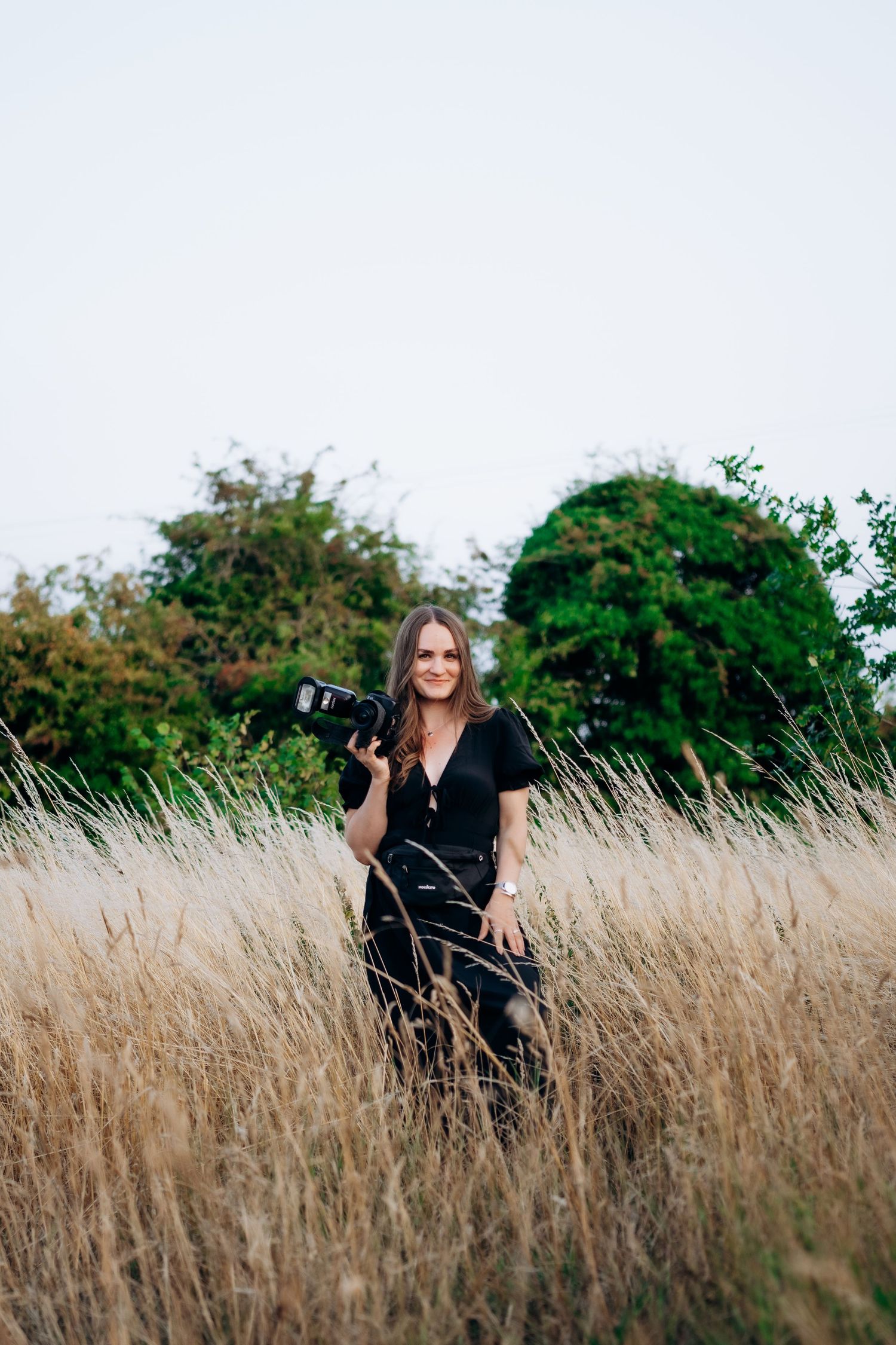 Professional cotswolds wedding photographer with camera crouches in tall golden grass while taking pictures in nature setting.
