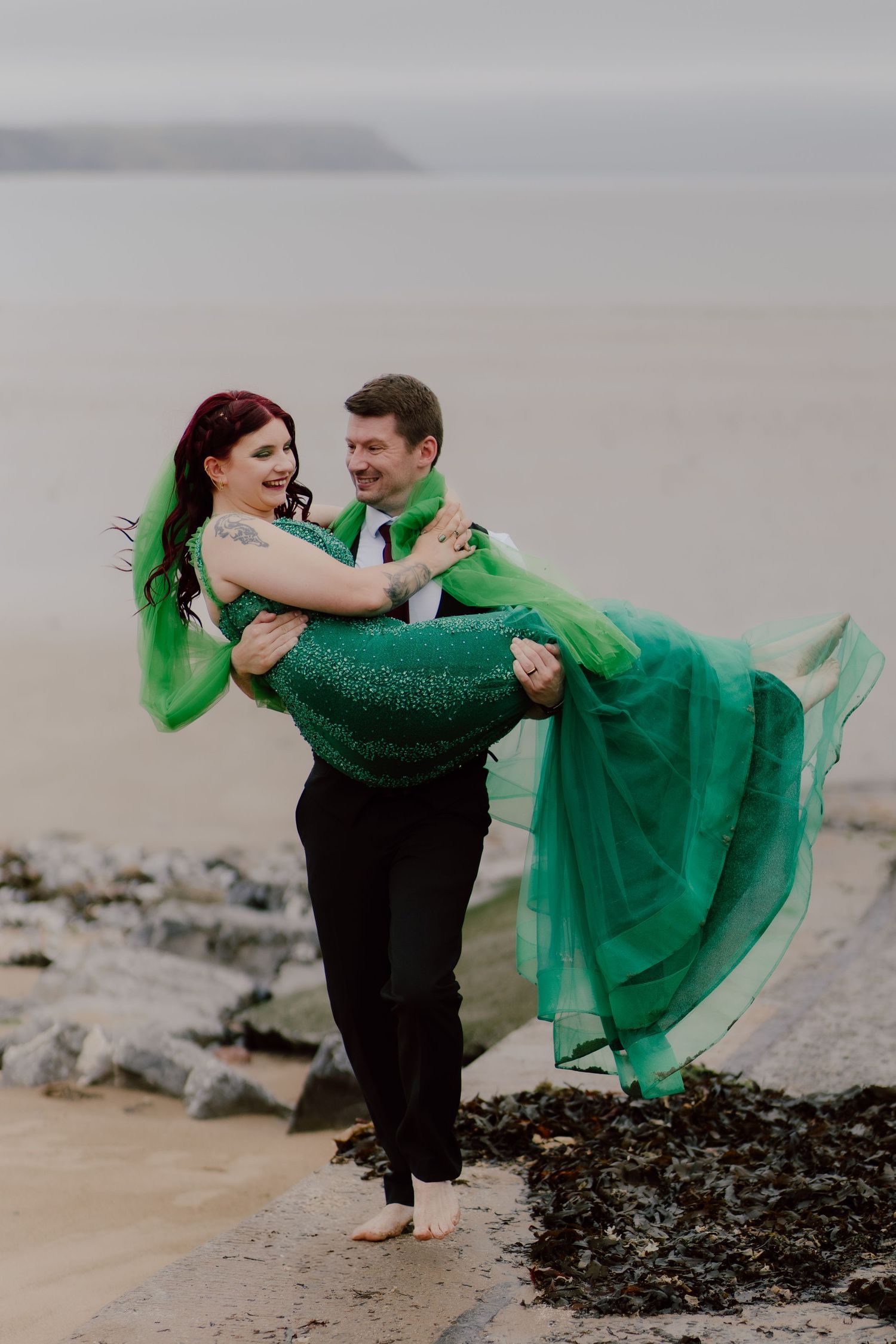 Romantic couple in green attire embrace on a misty beach, with flowing emerald dress creating dreamy coastal scene in Swansea.