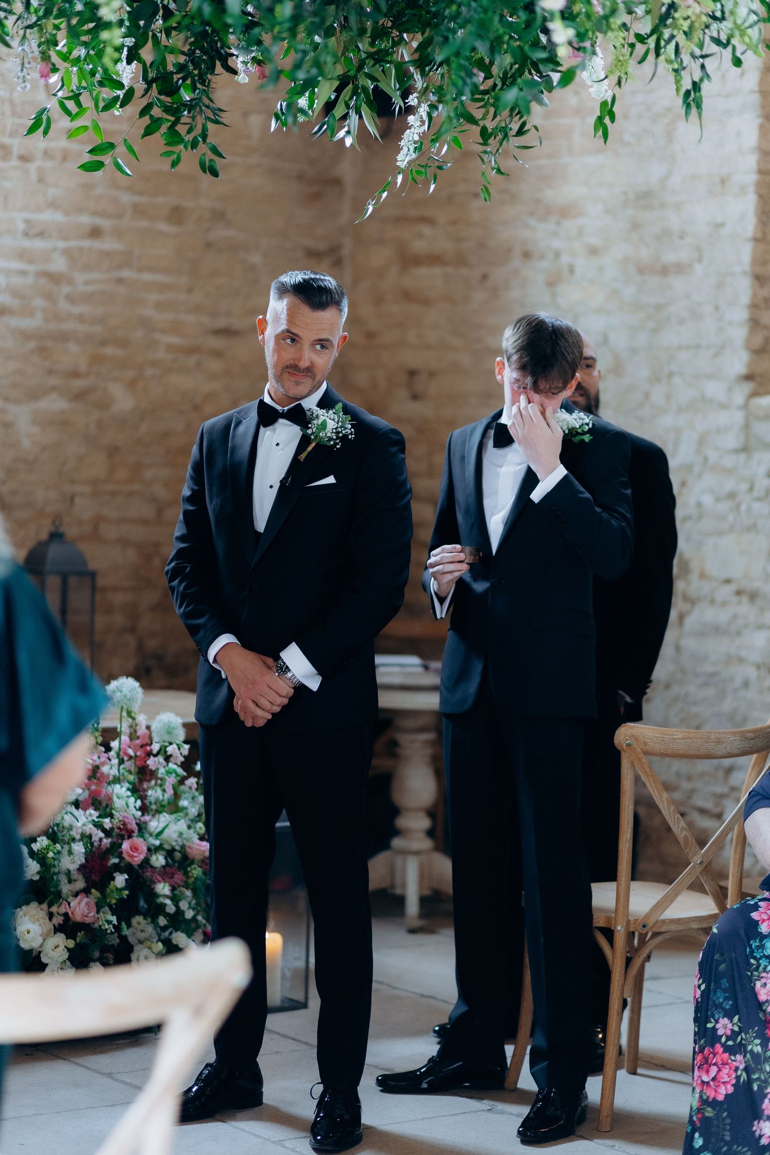 The groom and his best man in black tuxedos share an emotional moment at old gore wedding venue with brick walls and floral decorations.