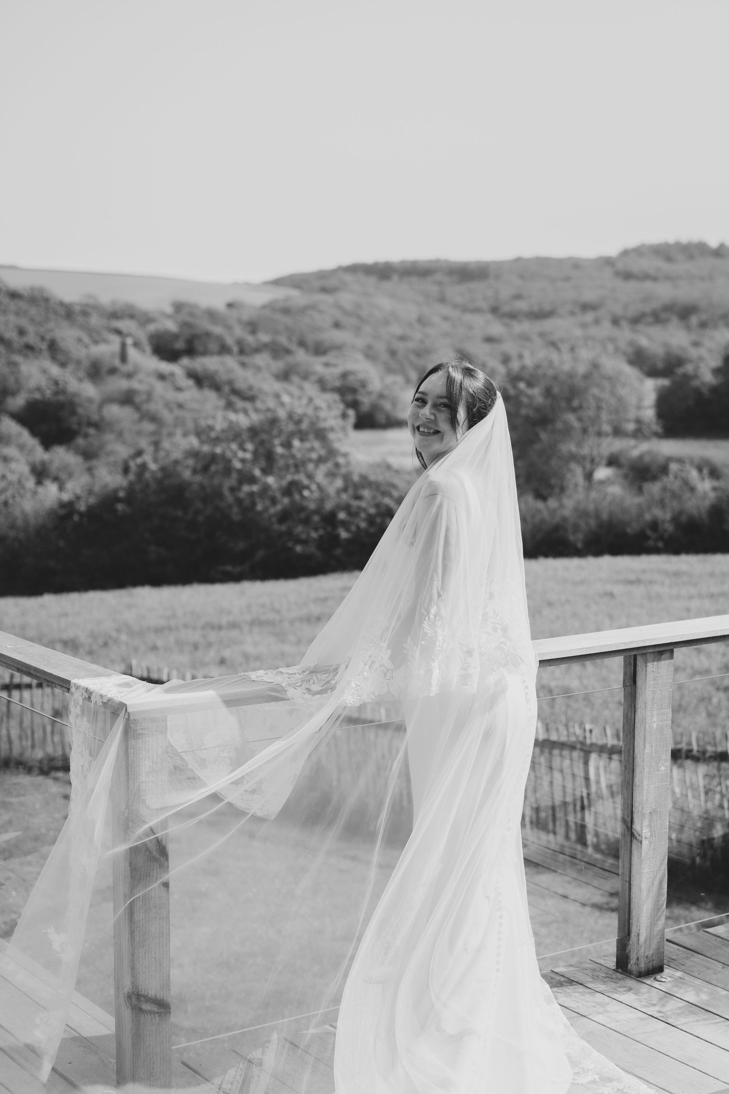 Black and white photo of a smiling bride in a flowing veil on a wooden deck with scenic countryside views.