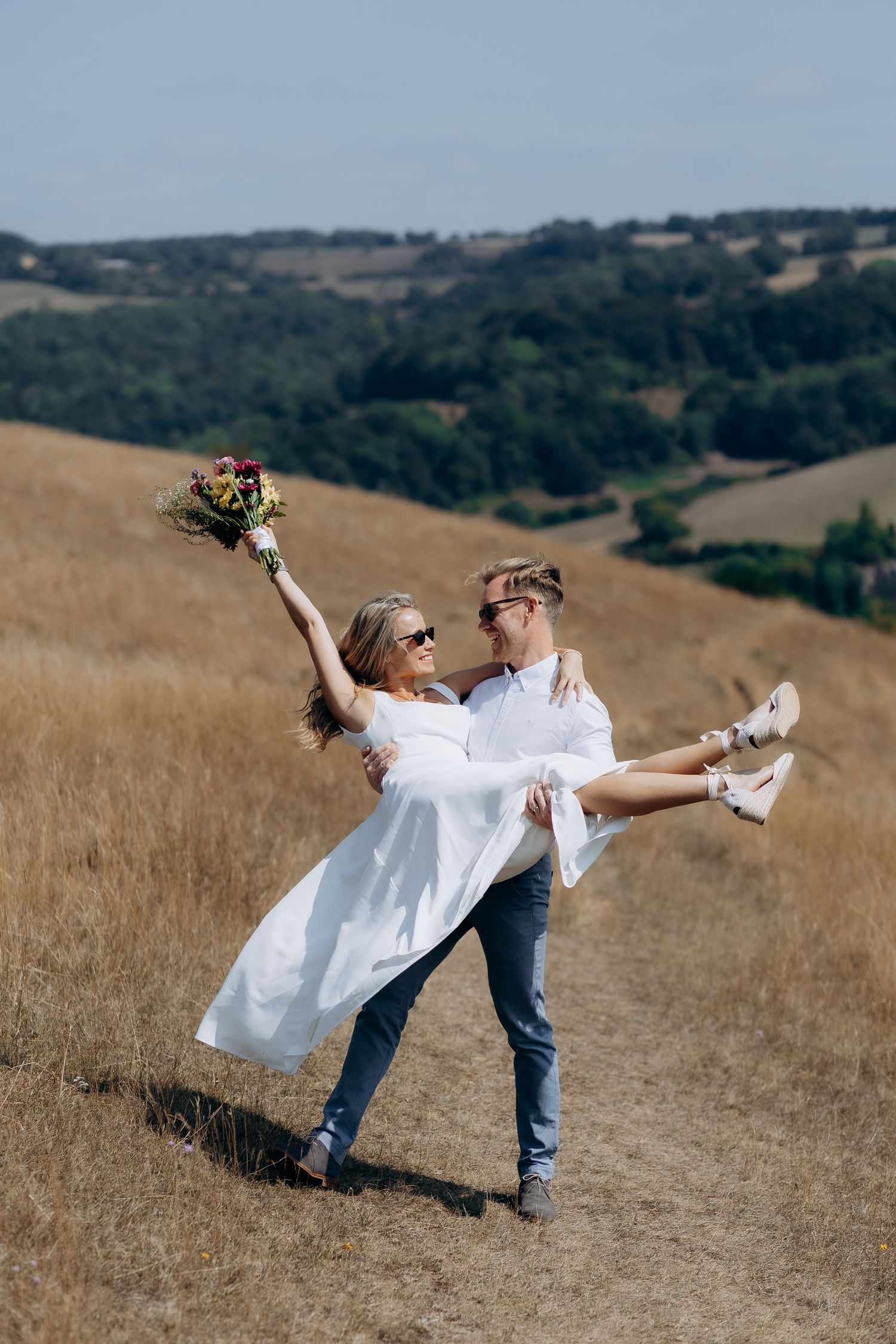 A couple embraces in a playful pose on a golden hillside with rolling hills in the background in the cotswolds.