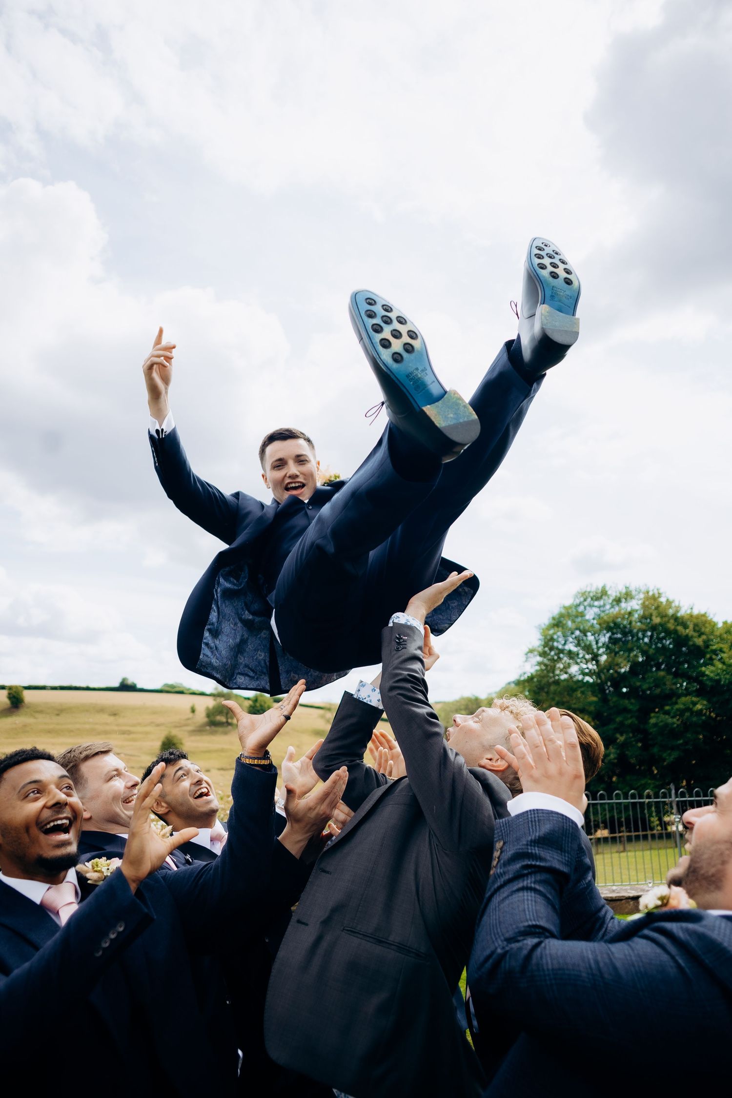 A group of groomsmen in dark suits toss the groom up in the air during wedding celebrations at Kingscote barn wedding venue.