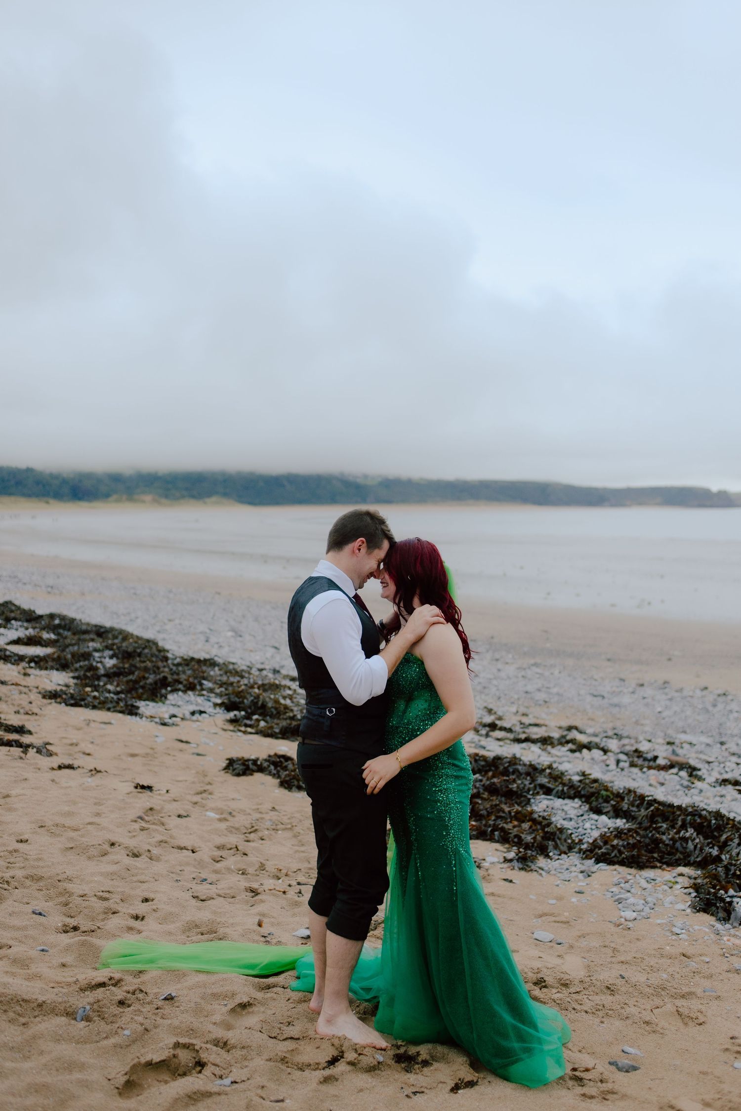 Romantic wedding couple embraces on a misty beach with rocky shore and overcast sky in the background captured by a uk wedding photographer.