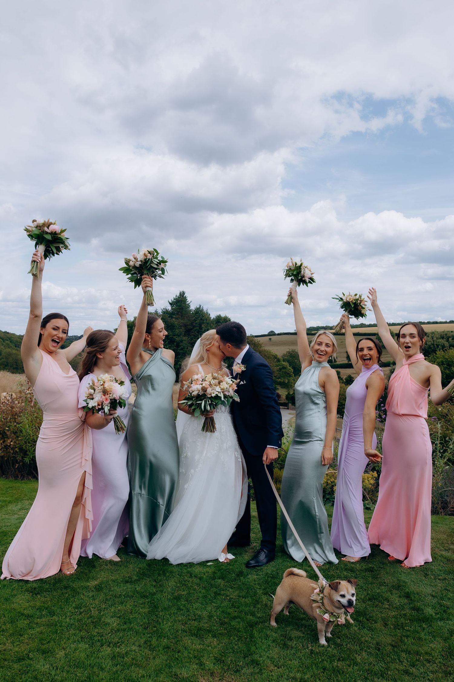 Wedding party celebrating and jumping joyfully at Kingscote barn wedding venue with colorful bridesmaid dresses and bouquets.