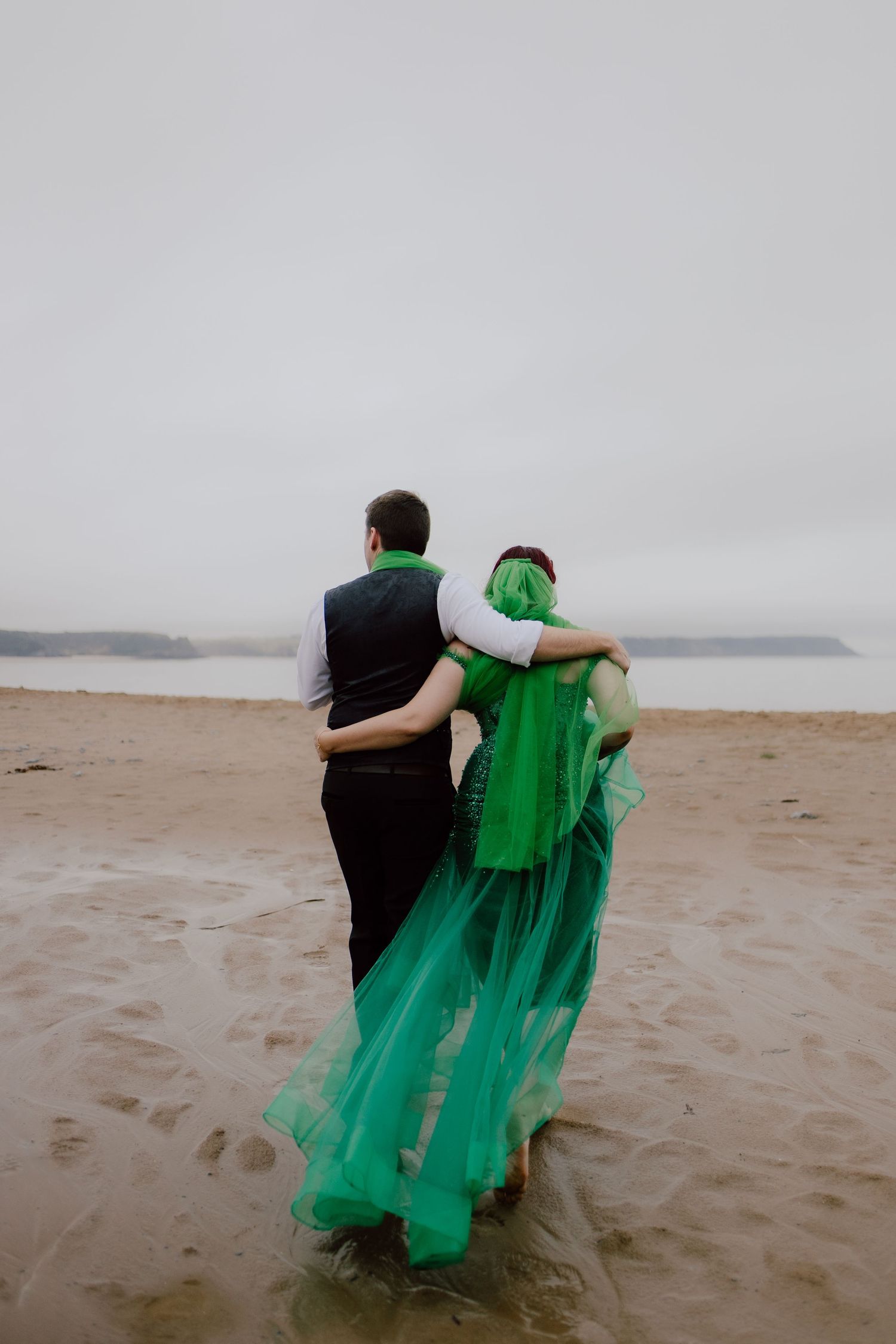 Two figures in black and emerald green attire walk together along a misty beach shoreline.