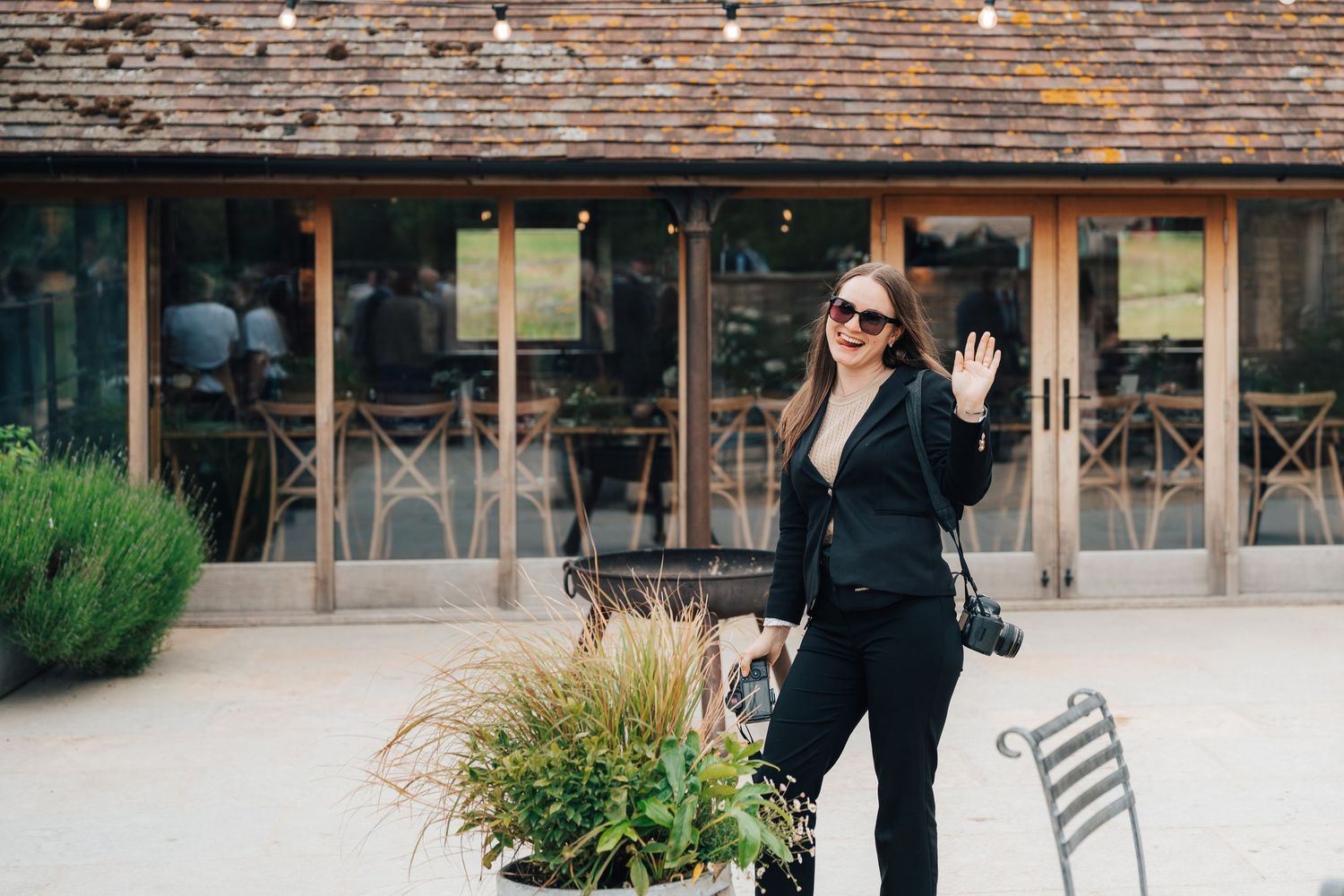 Professional wedding photographer in the cotswolds in an all-black outfit waves to the camera.