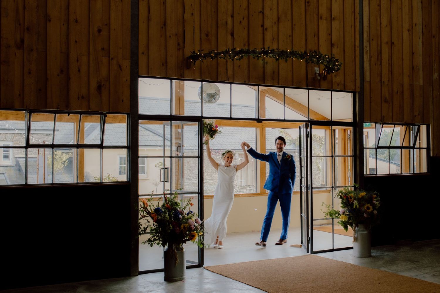 Bride and groom celebrate their grand entrance through elegant glass doors at a modern barn wedding venue in the cotswolds.