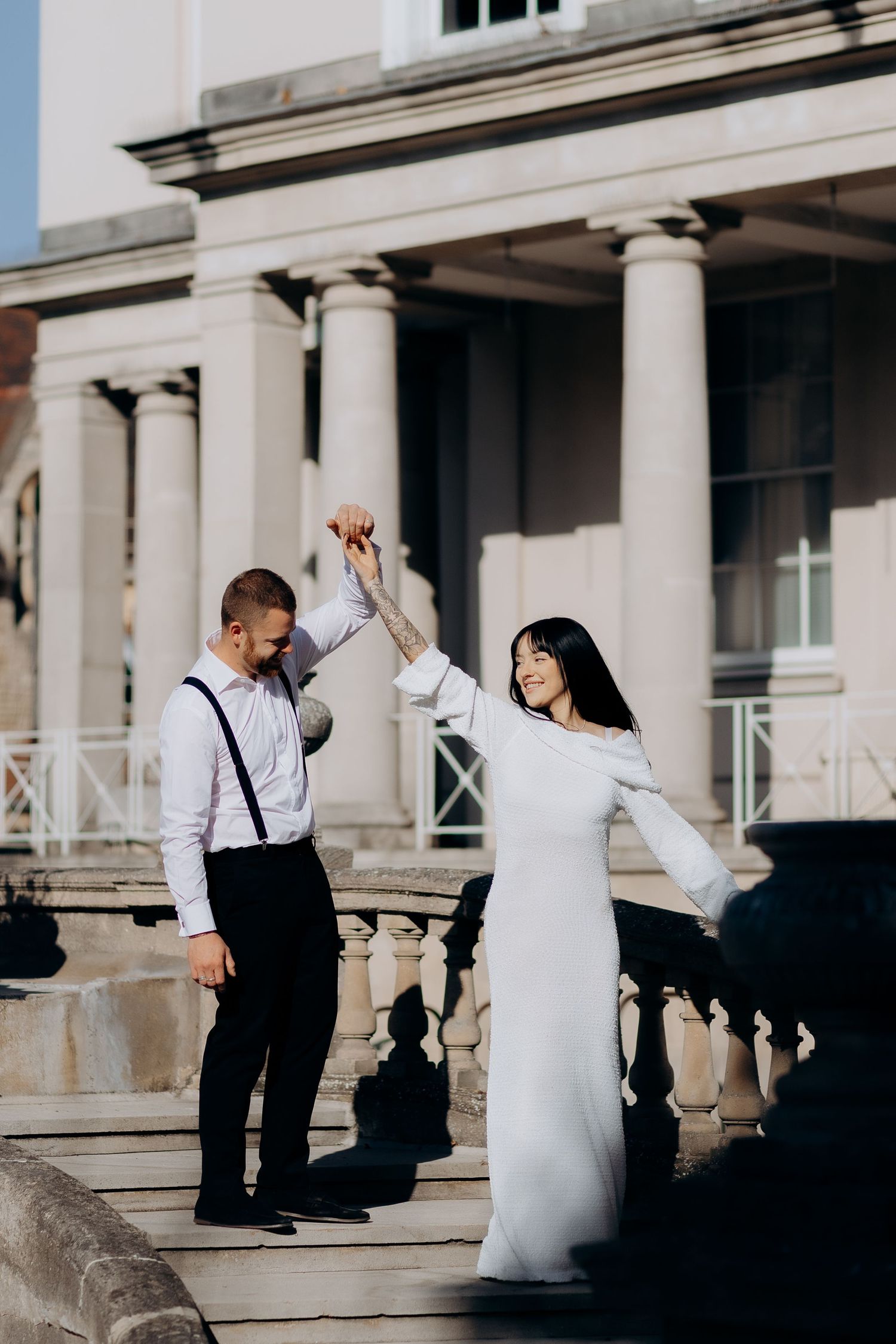Bride and groom stand on a grand columned balcony in cheltenham as the bride twirls while holding hands with groom in suspenders.