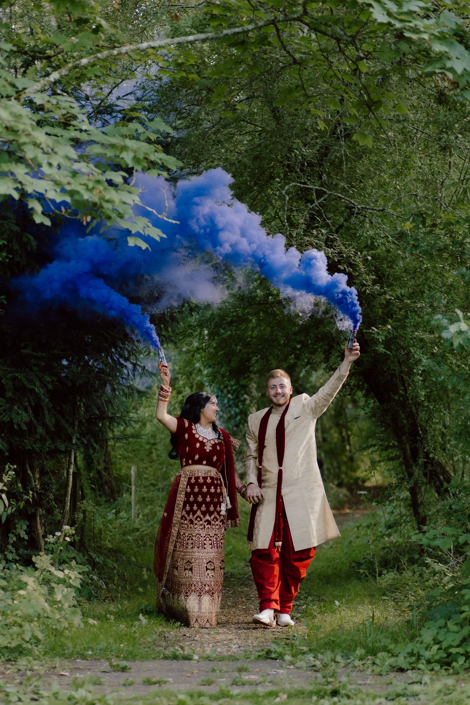 Two people in traditional Indian wedding attire hold blue smoke flares on a tree-lined path during a celebratory moment in the Cotswolds.