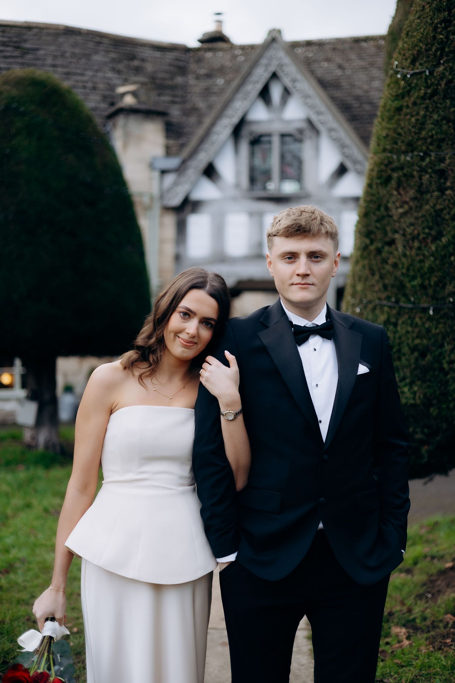 A bride and groom in wedding attire poses together at a garden wedding venue in the Cotswolds with traditional Cotswold stone architecture.
