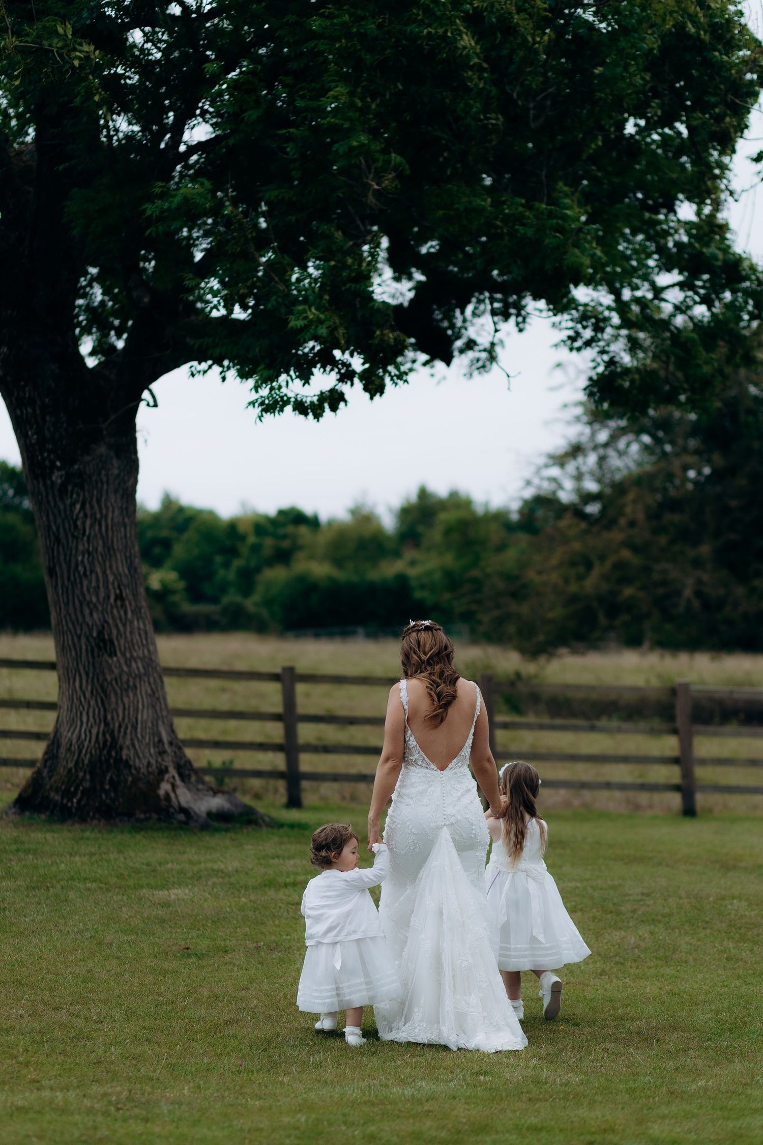A bride in a white gown walks hand in hand with two flower girls across a grassy field near a large tree and fence at Old Gore barn.